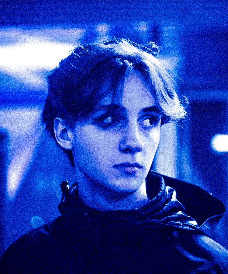 A young woman with short, wavy hair looking to the side in a dimly lit, blue-tinted environment.
