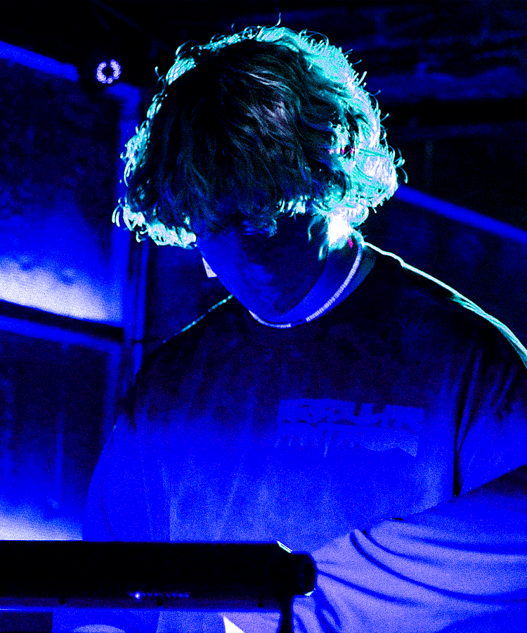 Person with curly hair wearing a dark shirt, illuminated by blue and purple lighting, looking down at electronic equipment.