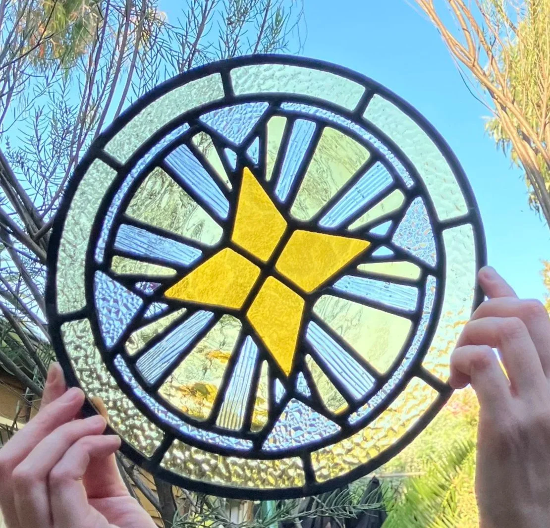 A stained glass circular art piece with a butterfly design in yellow and blue, held up against a clear sky with tree branches in the background.