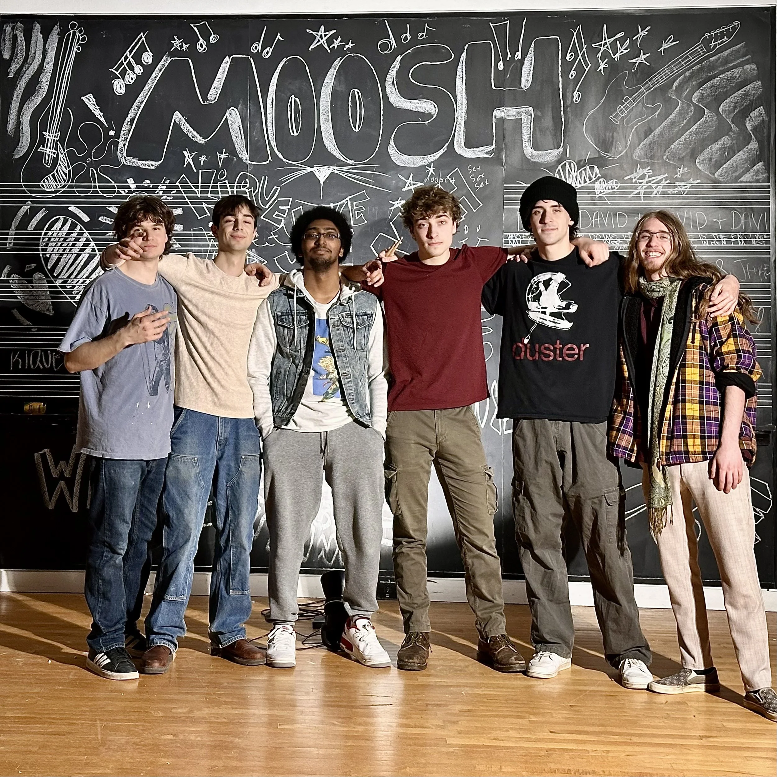 Six young men standing arm-in-arm on a wooden floor in front of a blackboard with musical and artistic drawings and the word 'MOSH' written at the top.