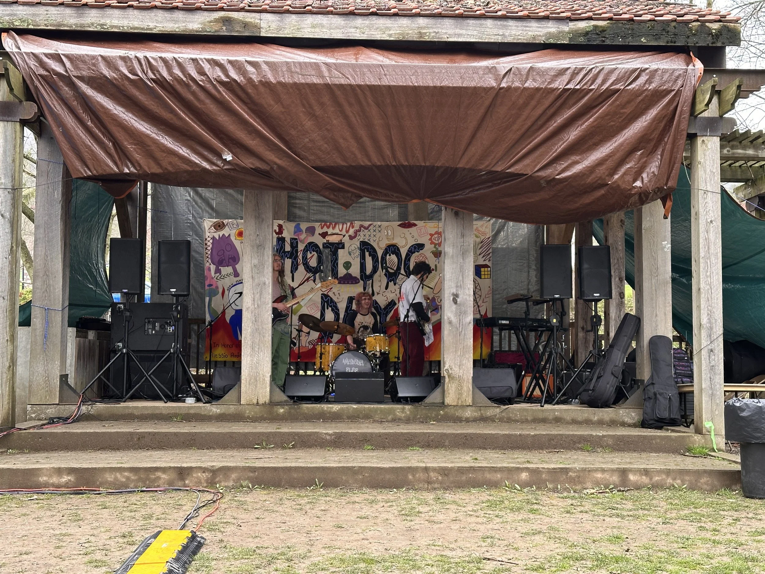 Band performing on an outdoor wooden stage with a colorful backdrop that says "Hot Dog". The band has four members, one playing drums, one on bass guitar, and two with electric guitars. Equipment including speakers, keyboards, and guitars is set up on stage.