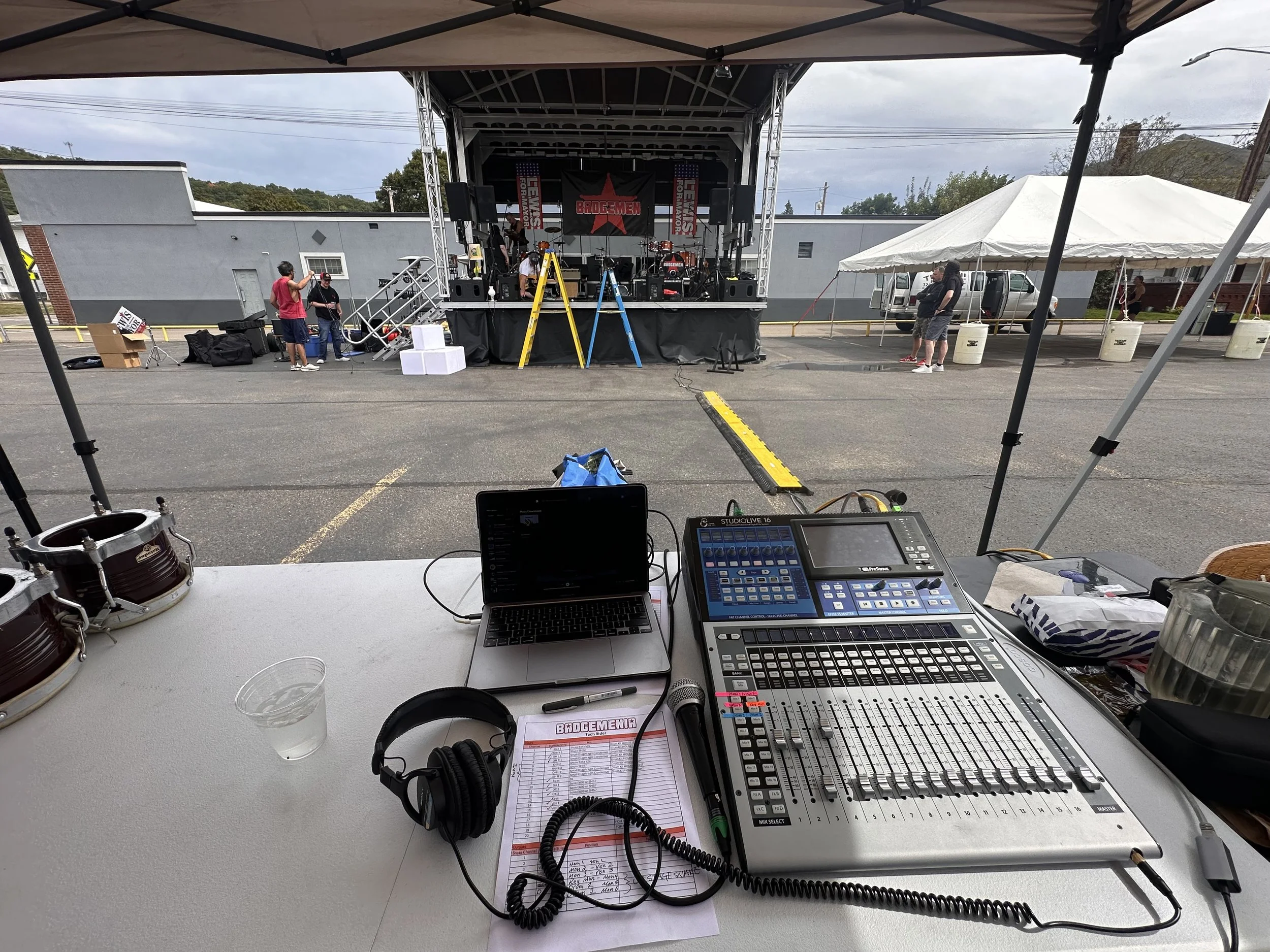 View of a stage setup with musical equipment, sound mixing console, laptop, headphones, and microphone in the foreground, in front of outdoor tents and a parking lot where a band is rehearsing before a performance.