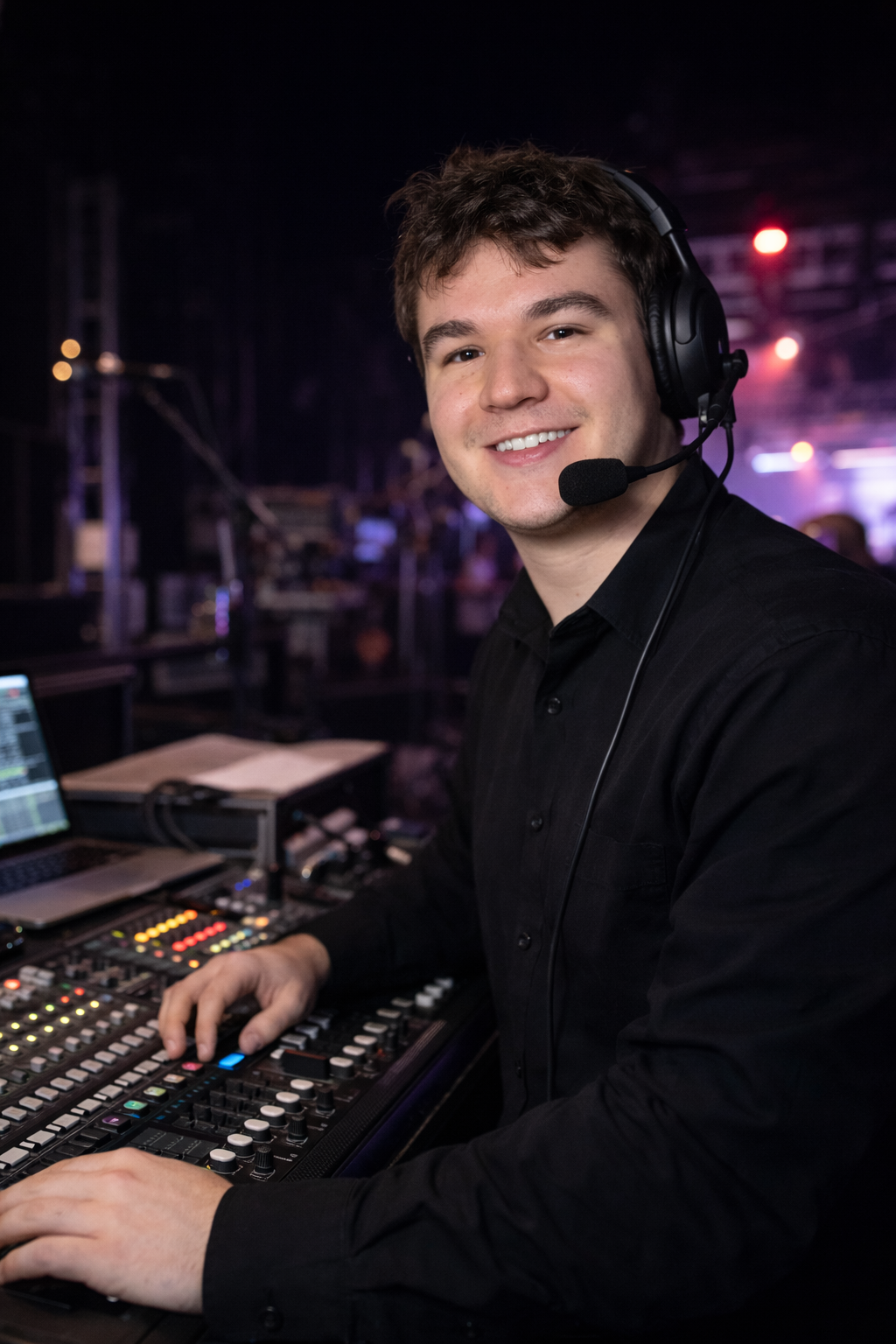A young man wearing a black shirt and a headset with a microphone, smiling at the camera, working as a DJ in a dimly lit venue with musical equipment and screens in front of him.