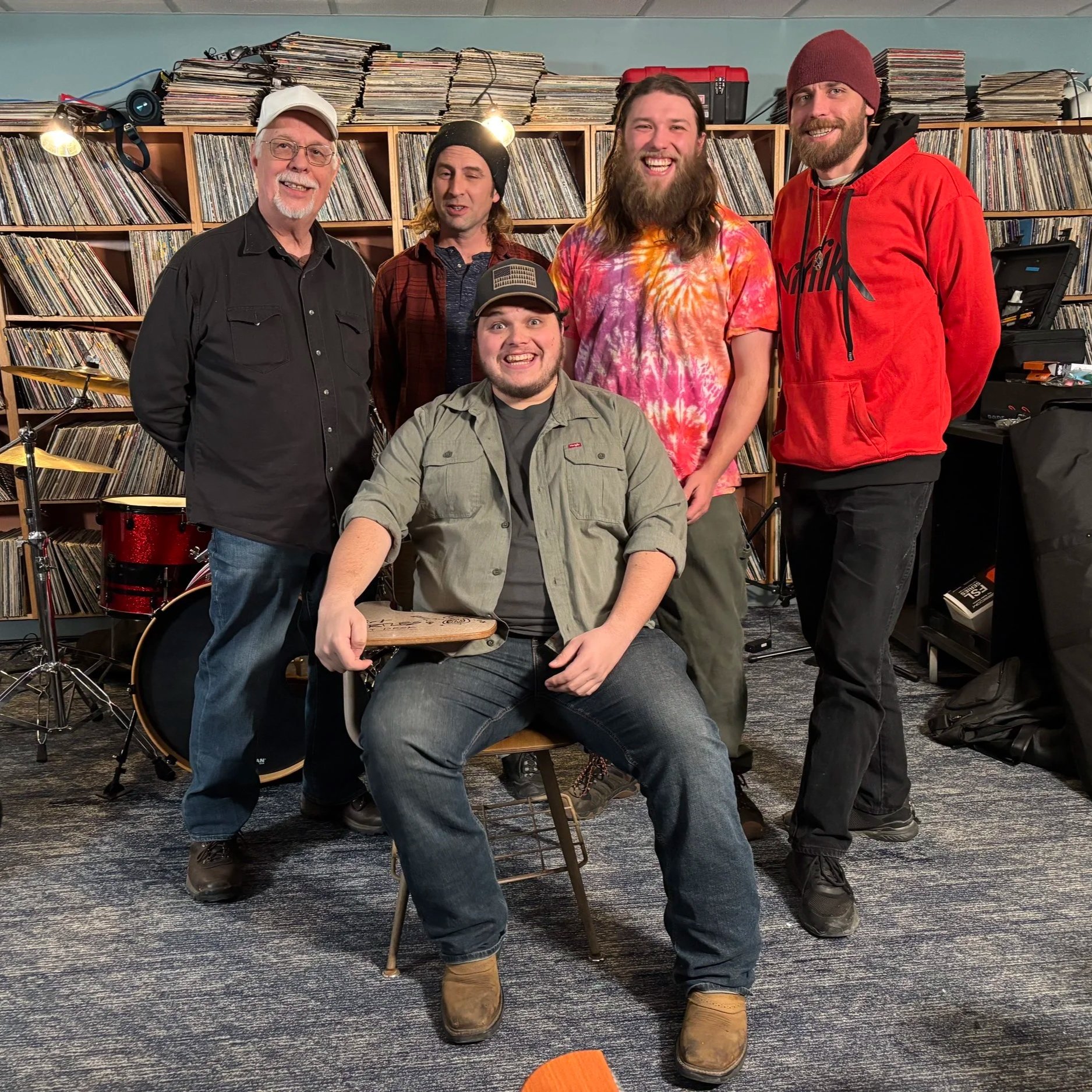 Group of five men in a music rehearsal space with shelves of vinyl records, musical instruments, and equipment.