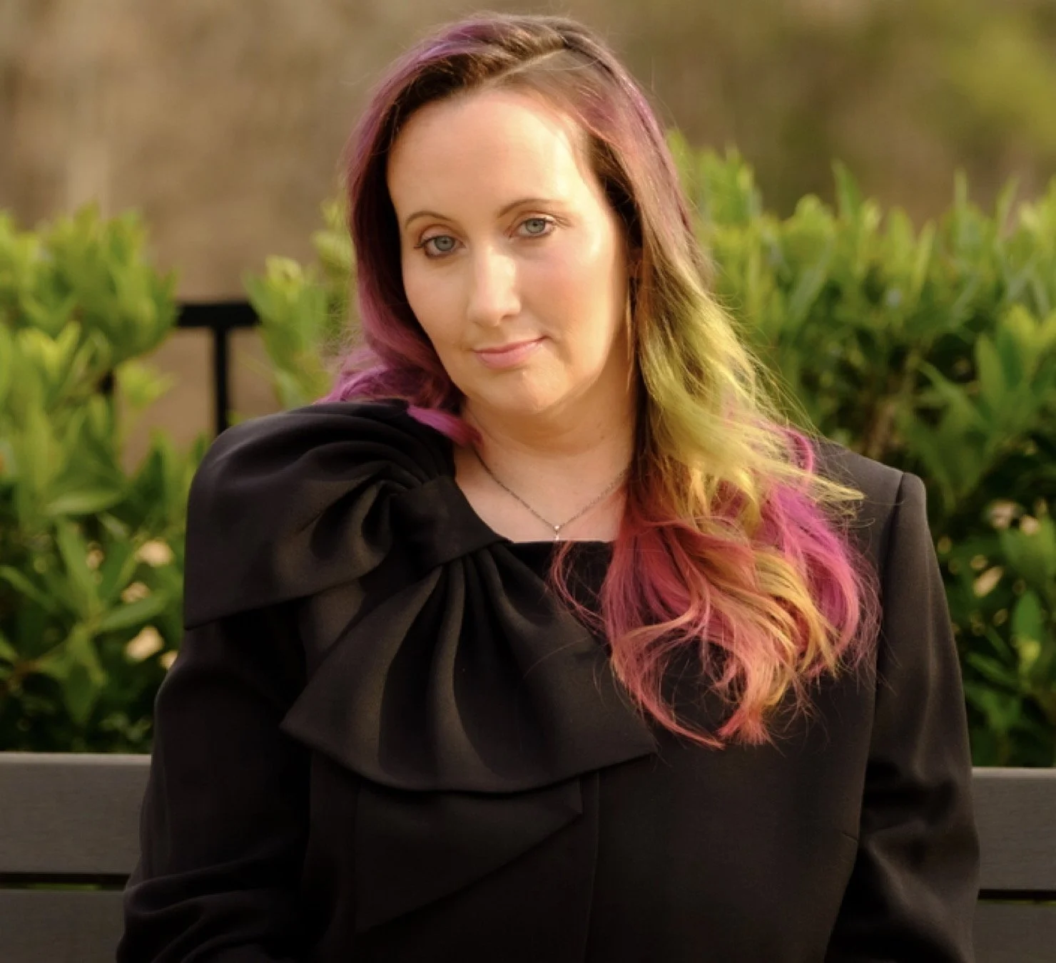 Commissioner-at-large candidate Heather Morrow is front and center, in a headshot. She has mostly pink hair with green in it. She has blue eyes and fair skin and is sitting in the sun. She is wearing a black top with a large black bow on the shoulder