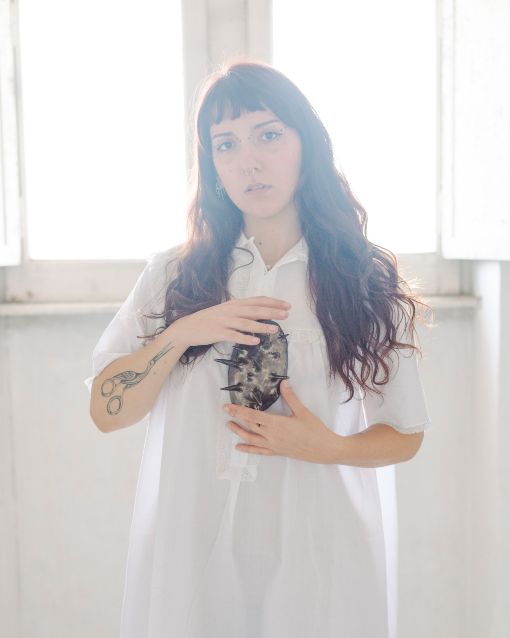 Donna con lunghi capelli ricci, tatuaggi e piercing, indossa un abito bianco e tiene un oggetto in ceramica artigianale spinoso di fronte al petto, con luce brillante sullo sfondo di una finestra.