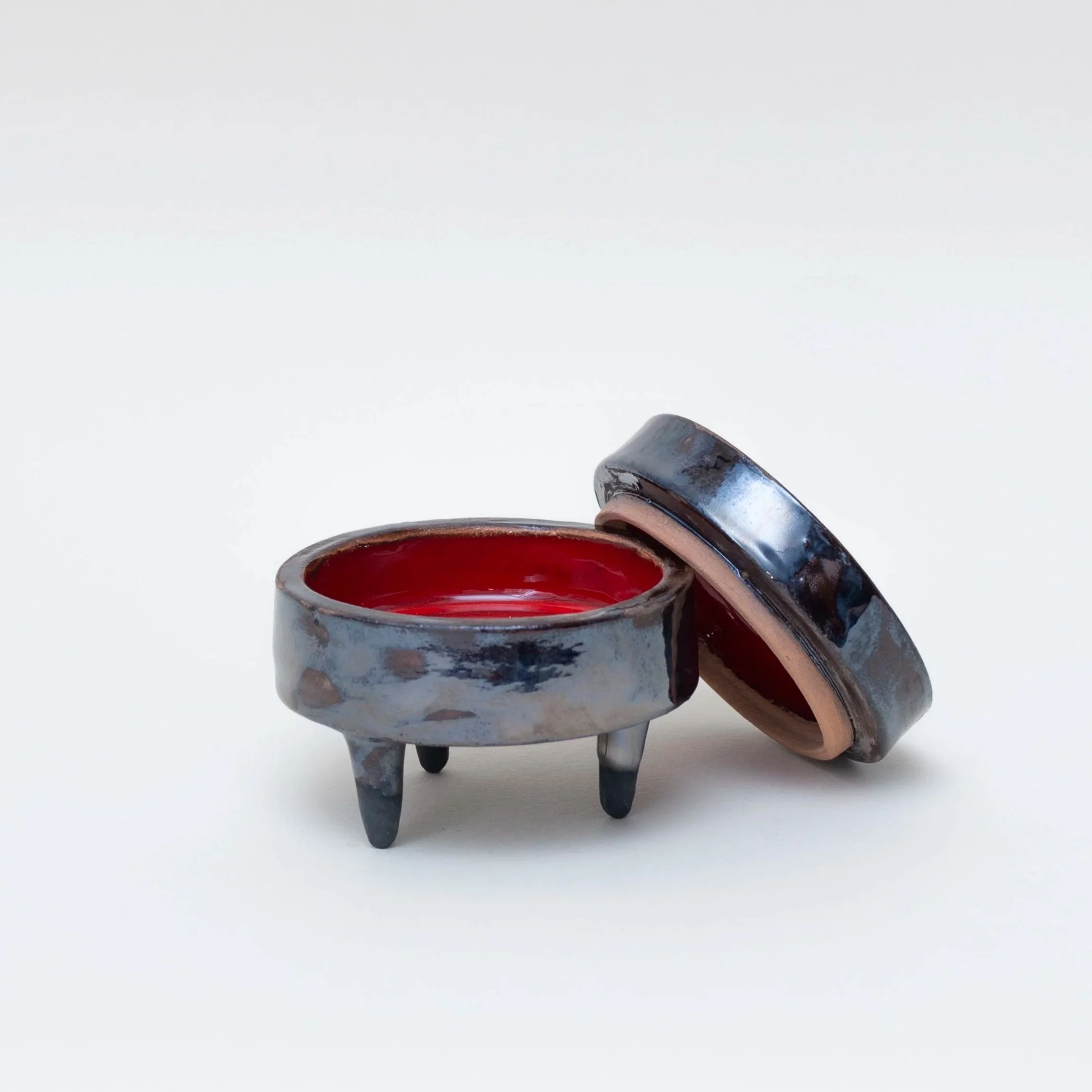 Contenitore di ceramica artigianale con interno rosso e piedini, uno con coperchio, posizionati su uno sfondo bianco.