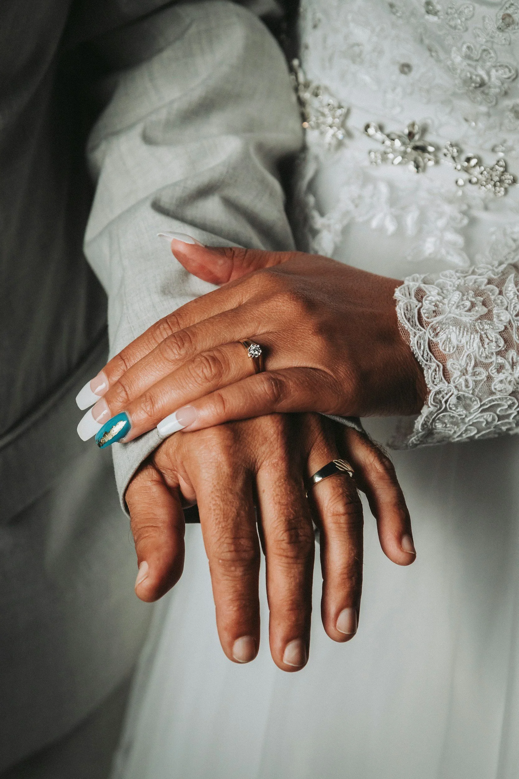 Close-up of a couple's hands with wedding rings, one hand resting on top of the other, with the woman's hand showing a blue nail with a gold design, and part of her lace wedding dress visible.