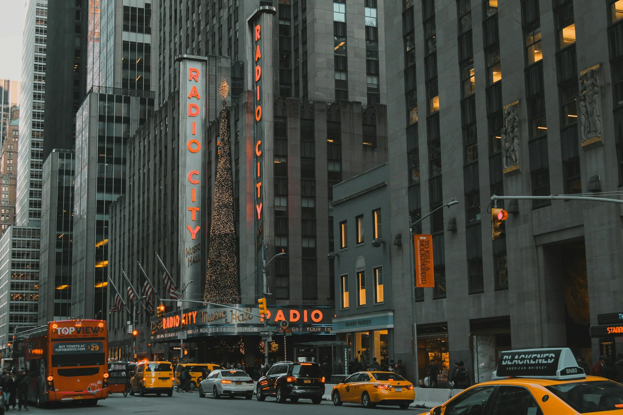 Nighttime street scene in New York City with a large Radio City Music Hall marquee, neon signs, yellow taxis, and pedestrians.