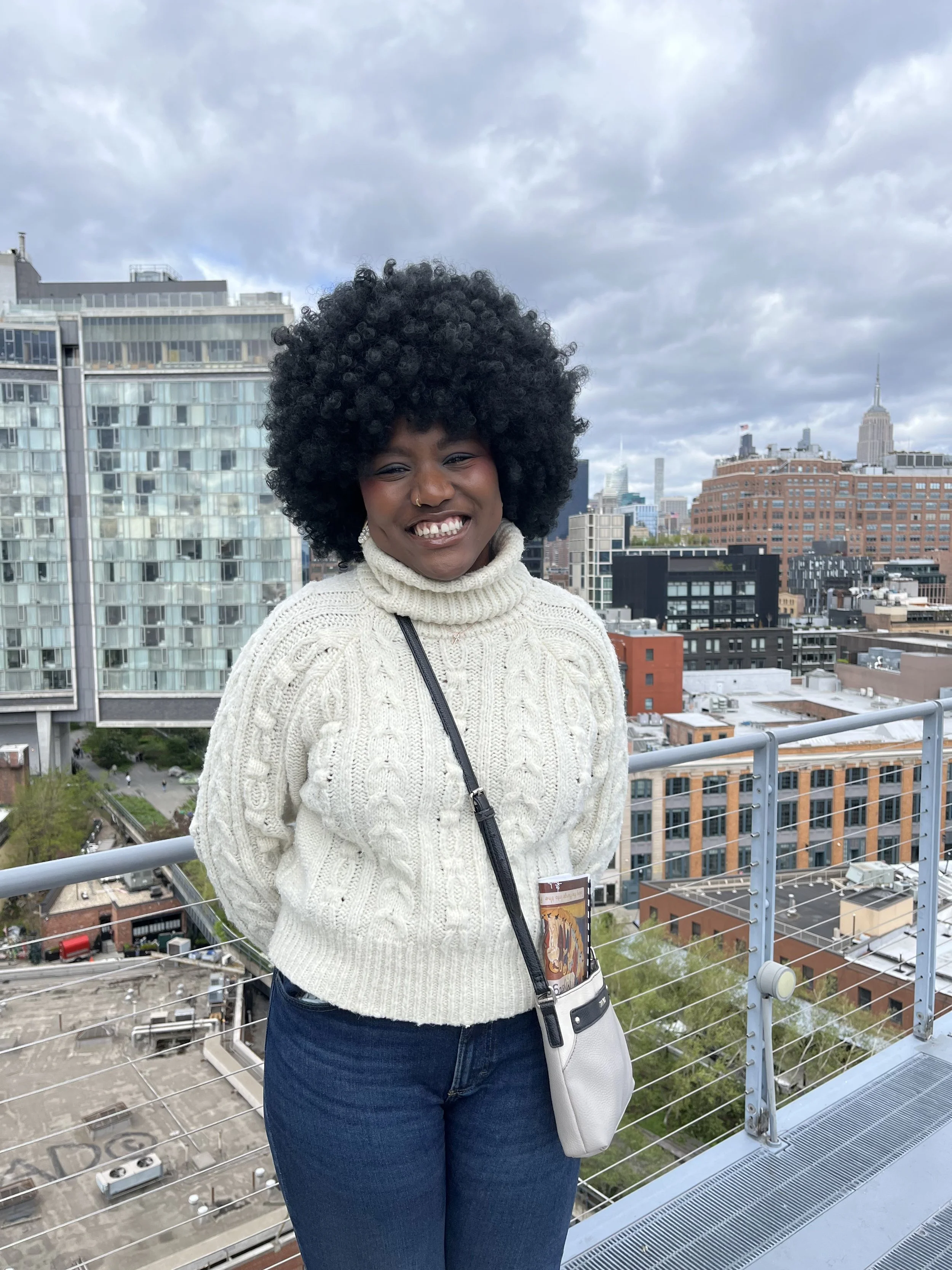 A woman with dark skin and a large curly afro hairstyle standing on a rooftop with a cityscape background. She is smiling, wearing a cream-colored cable knit turtleneck sweater, blue jeans, and has a crossbody bag.