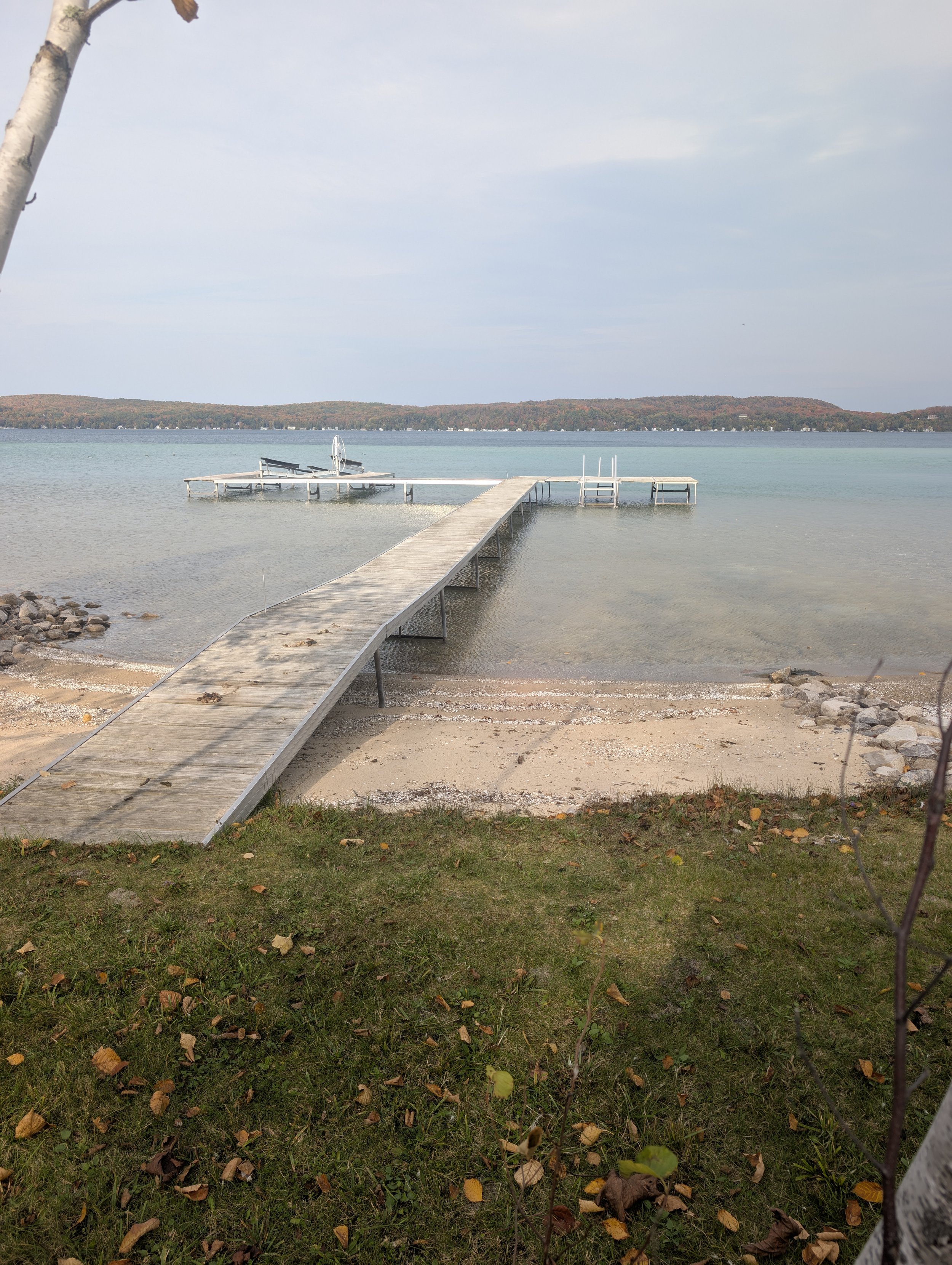 A wooden dock extending into a lake with small boat docks at the end, surrounded by a beach and grassy area with fallen leaves, and hills in the distance under a cloudy sky.