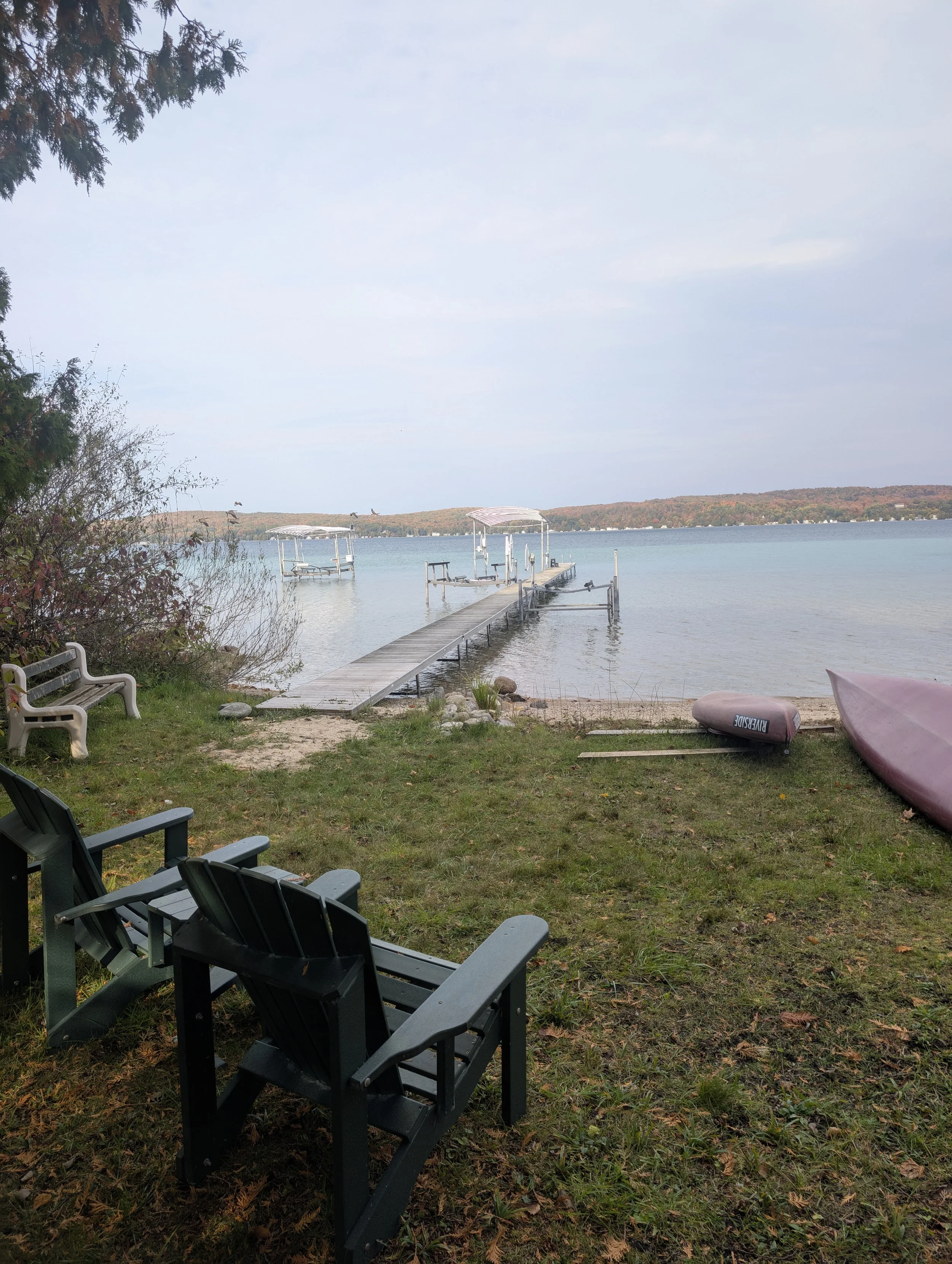 A lakeside scene with a grassy area featuring three Adirondack chairs, a small beach with a dock extending into the water, kayaks on the shore, and trees and distant hills in the background.