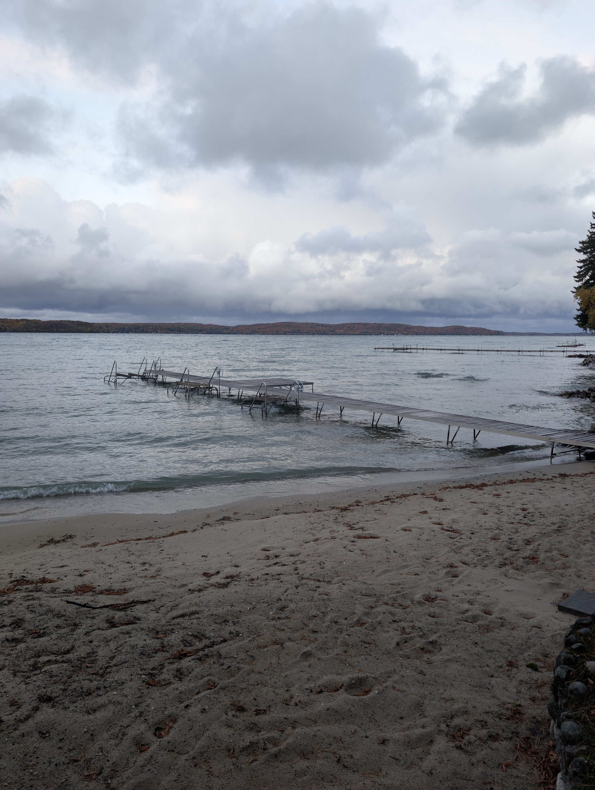 Overcast sky with a lake, a dock extending into the water, sandy beach in foreground, and distant land with trees.