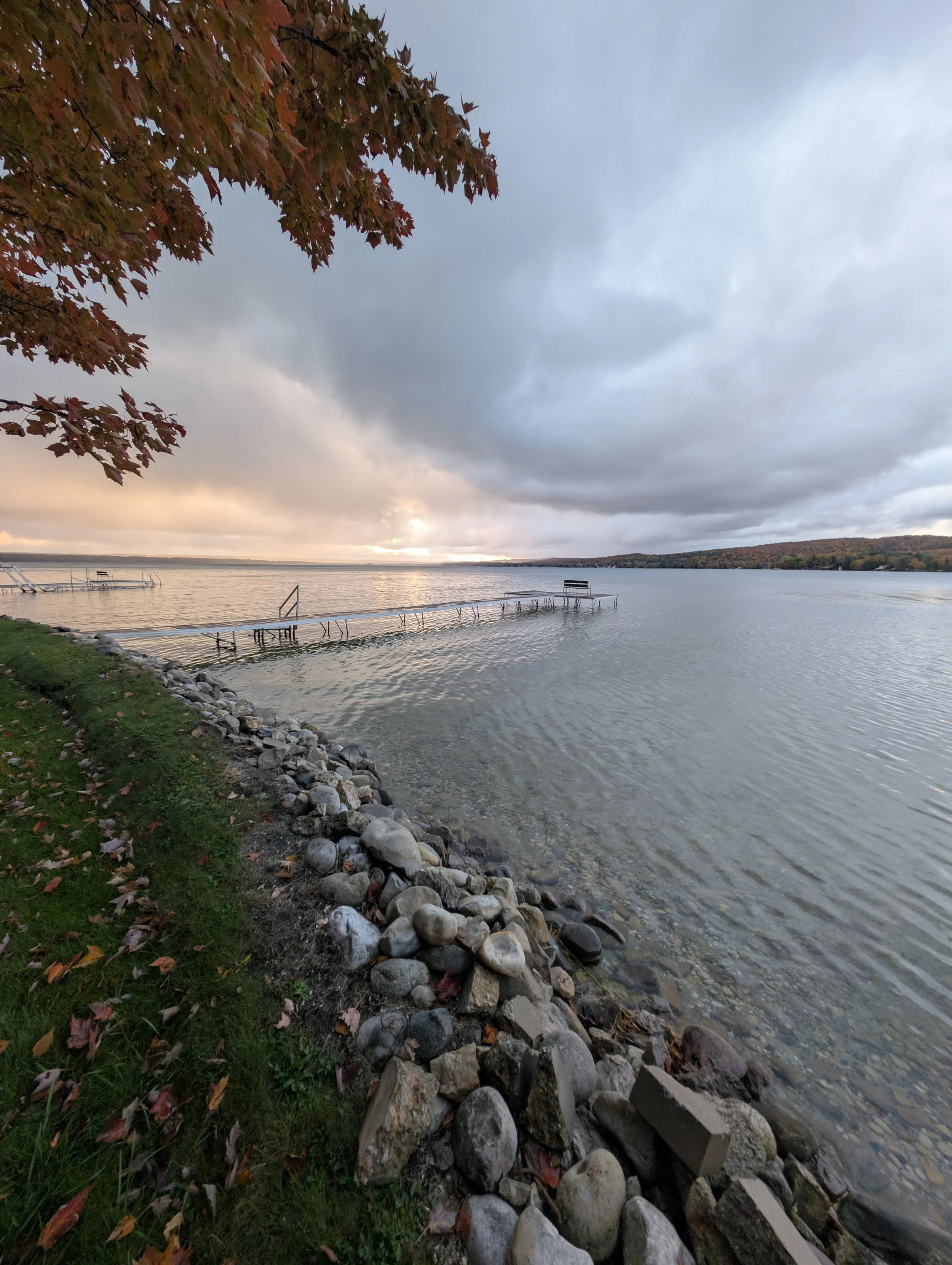 Calm lake with lapping water, stone-lined shoreline, dock, benches, and partially cloudy sky during sunset, with autumn foliage in the foreground.