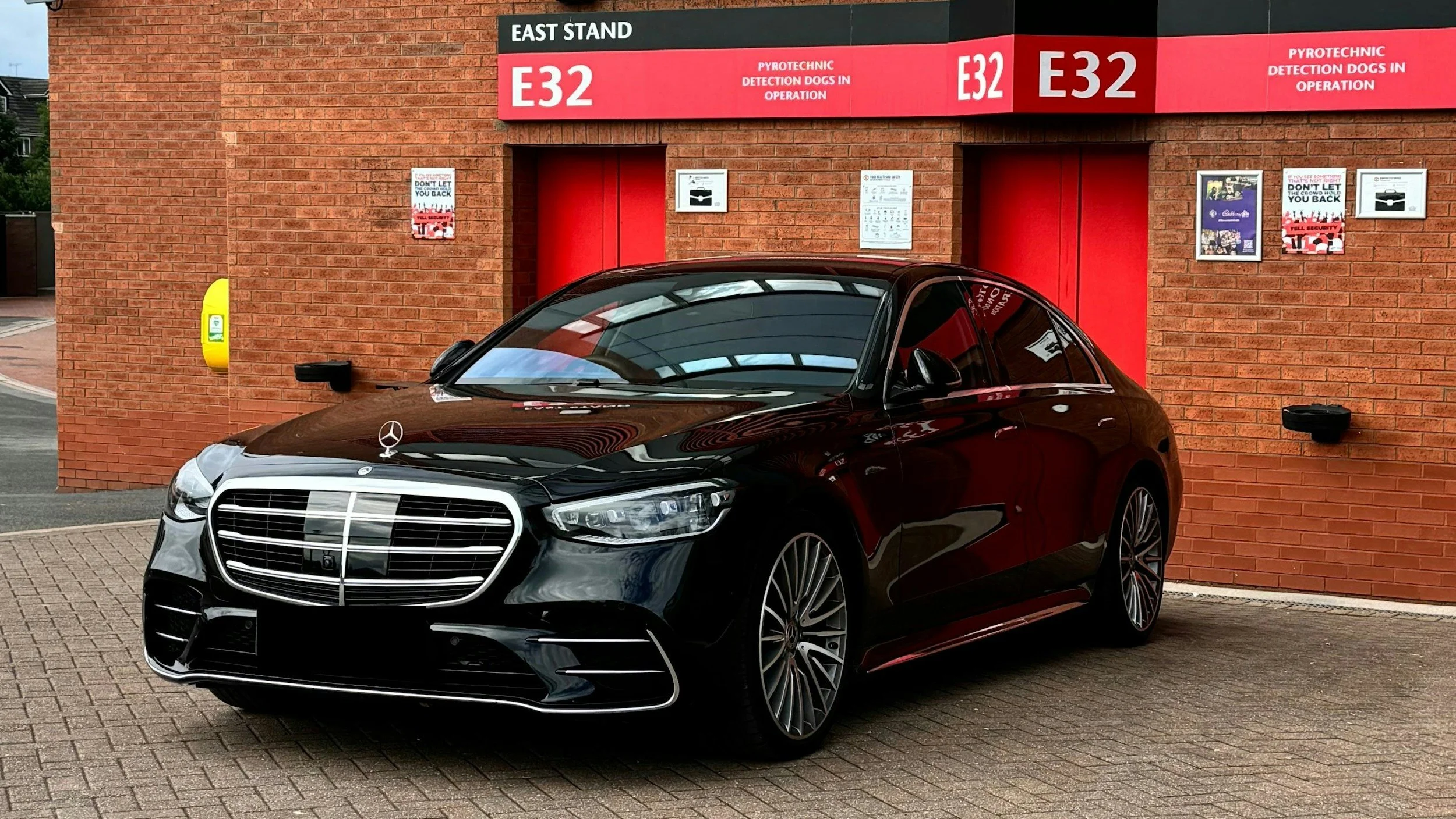 Black Mercedes-Benz car parked outside a red brick building with signs for East Stand and pyrotechnic detection dogs in operation