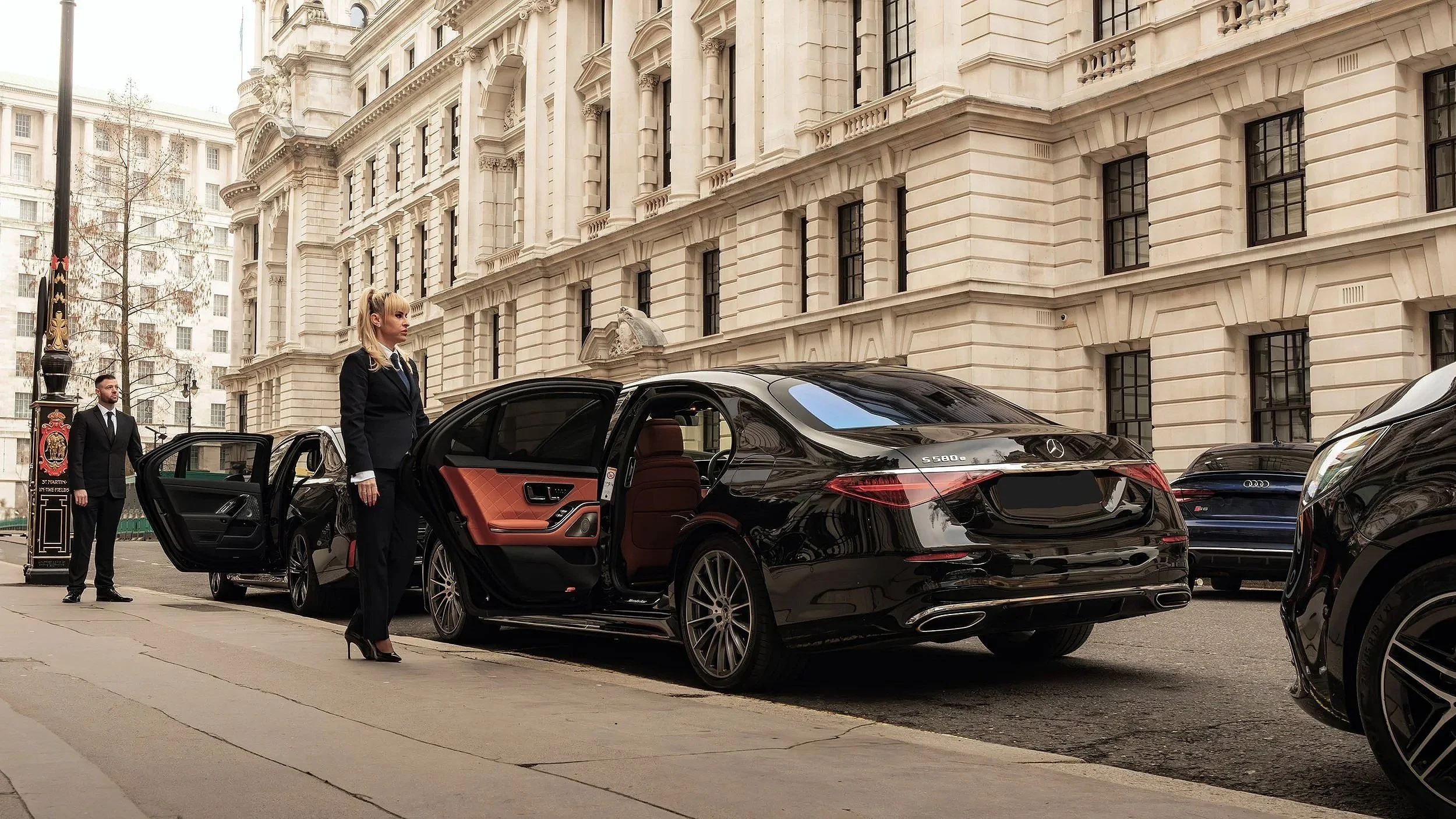 A woman in a black suit and high heels stands outside of a black luxury sedan on a city street, with a man in a suit and another luxury vehicle in the background, in front of historic white buildings.