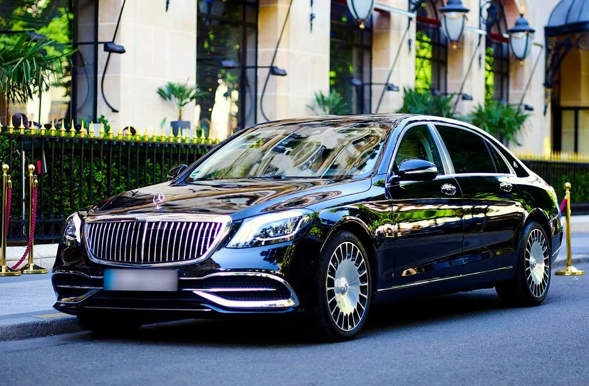 A black luxury Mercedes-Benz sedan parked on a city street with a building and greenery in the background.