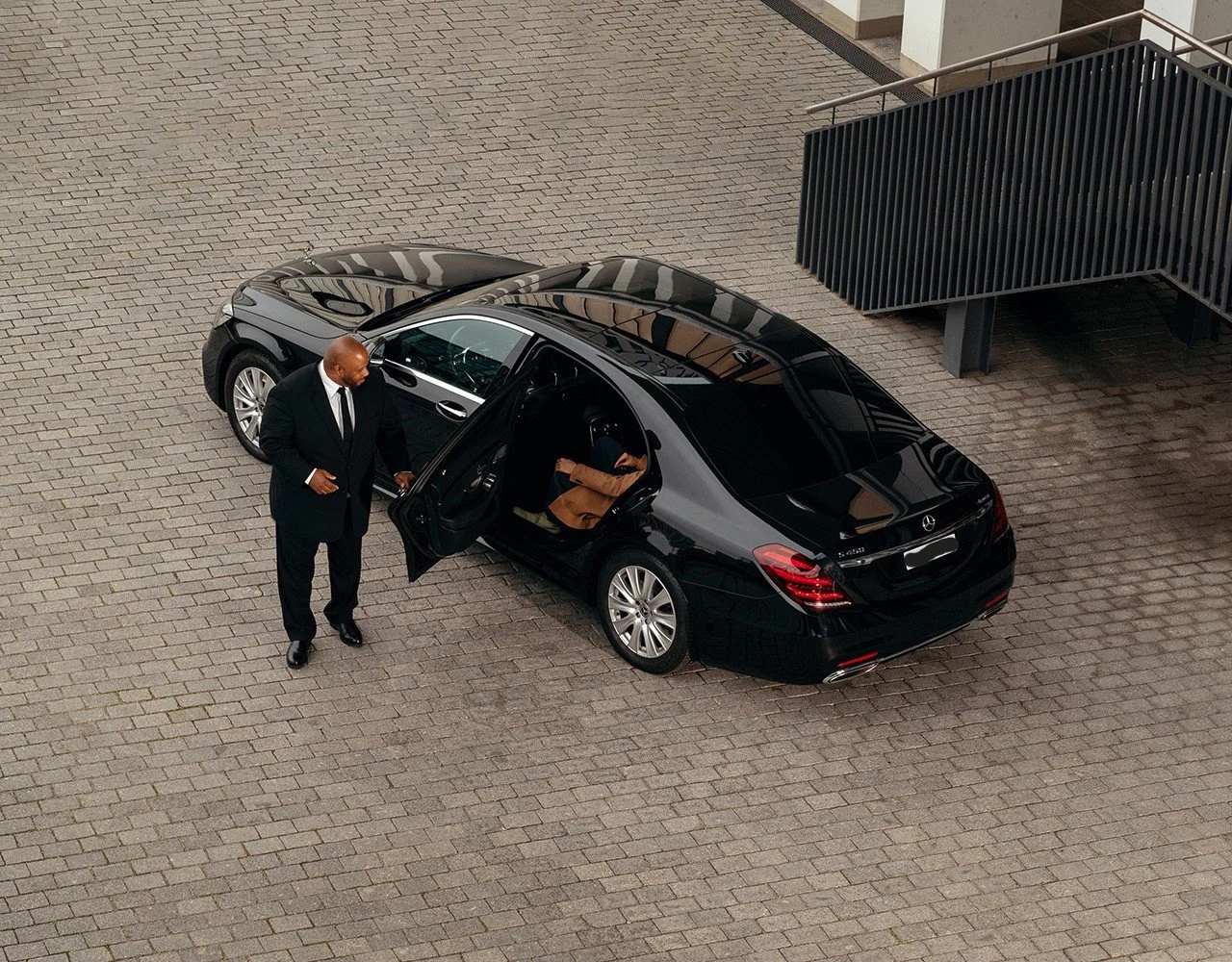 A man in a black suit standing outside a black luxury sedan, with a child sitting in the back seat. The setting appears to be a paved area near a building with a staircase.
