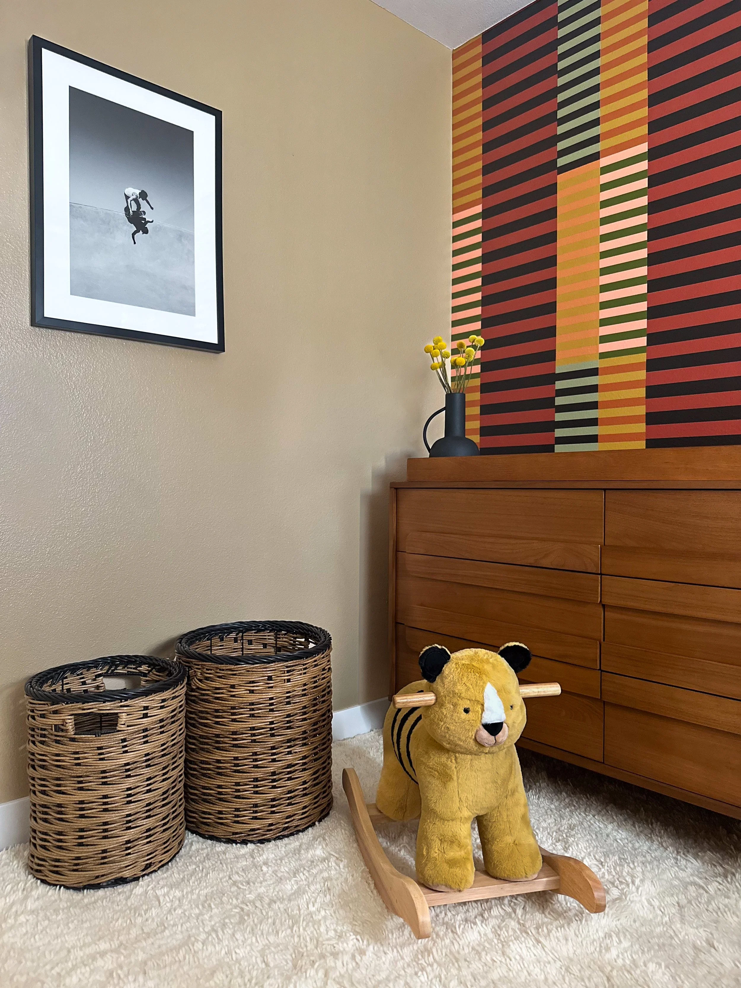 A cozy nursery corner with a rocking lion plush toy, two woven baskets, a wooden dresser, a black vase with yellow flowers, and framed monochrome artwork on the wall. The wall behind the dresser features colorful striped wallpaper.