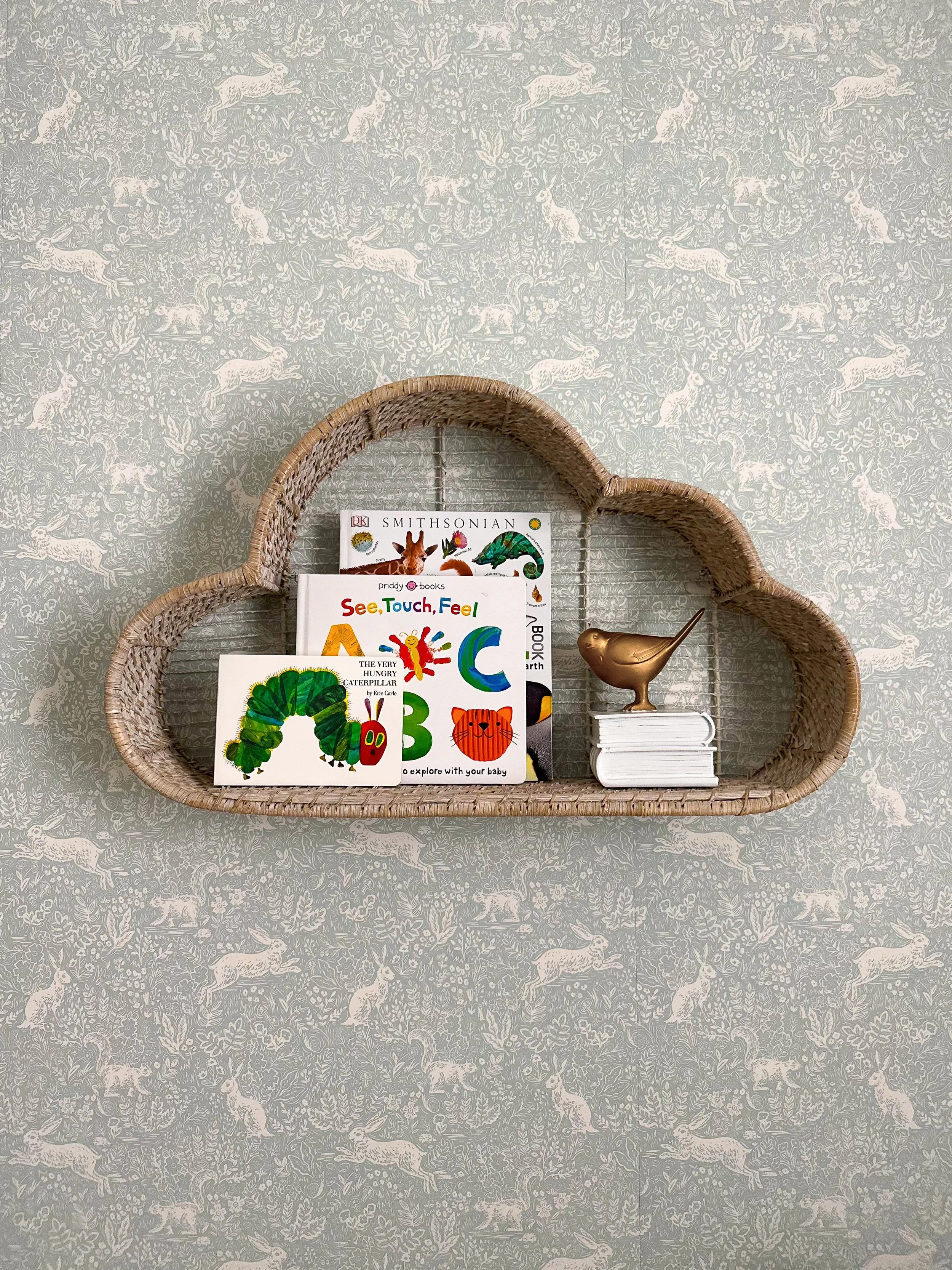 Wicker cloud-shaped shelf holding children's books and a gold bird figurine on a patterned wallpaper background.
