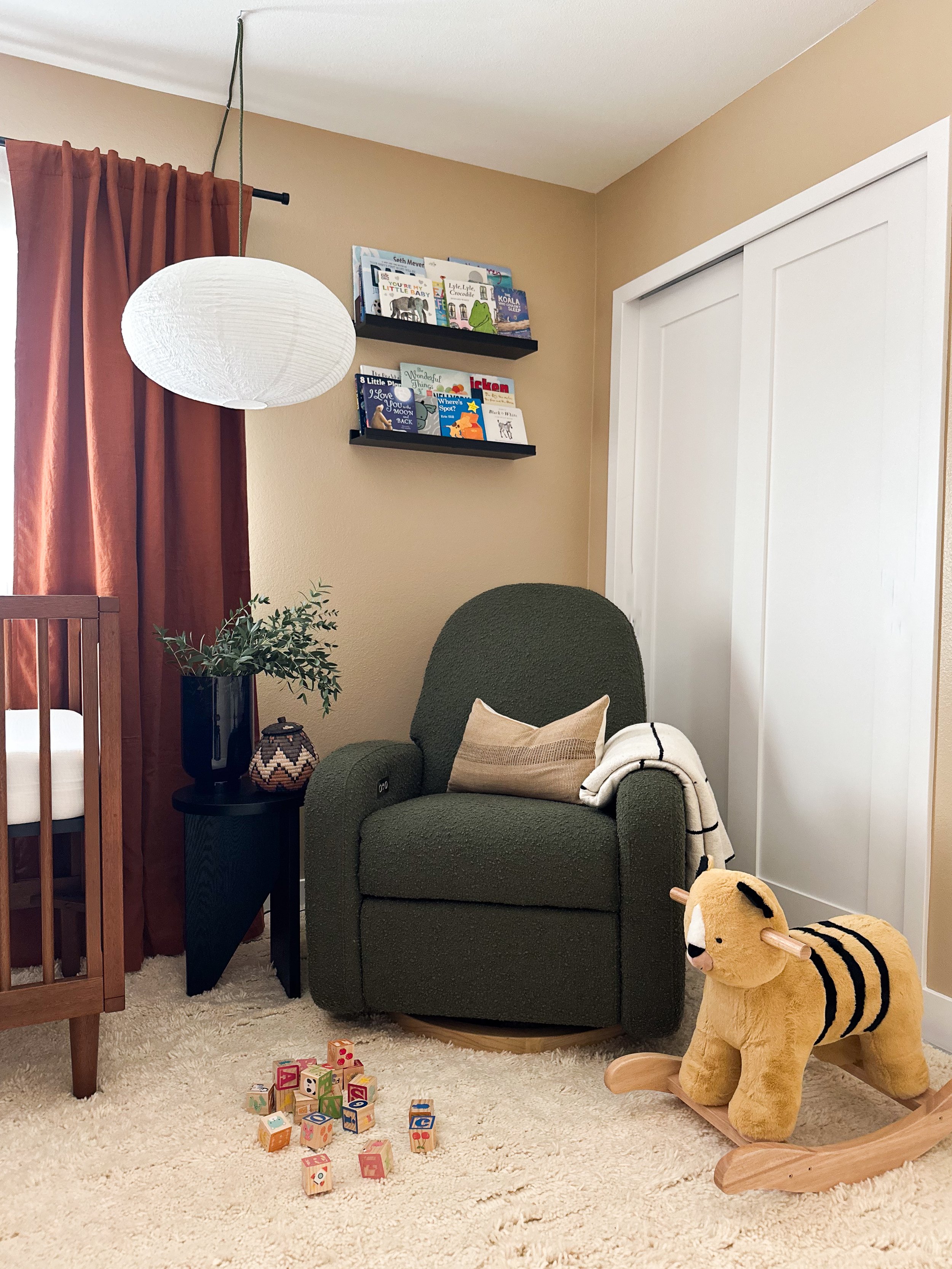 A cozy corner of a child's room with a green armchair, a beige pillow, a blanket, a plush tiger rocking horse, a small black side table with a plant and decorative vase, wooden colorful alphabet blocks, a white ceiling lamp, and a wall with shelves of children's books, beige walls, brown curtains, and a white closet door.
