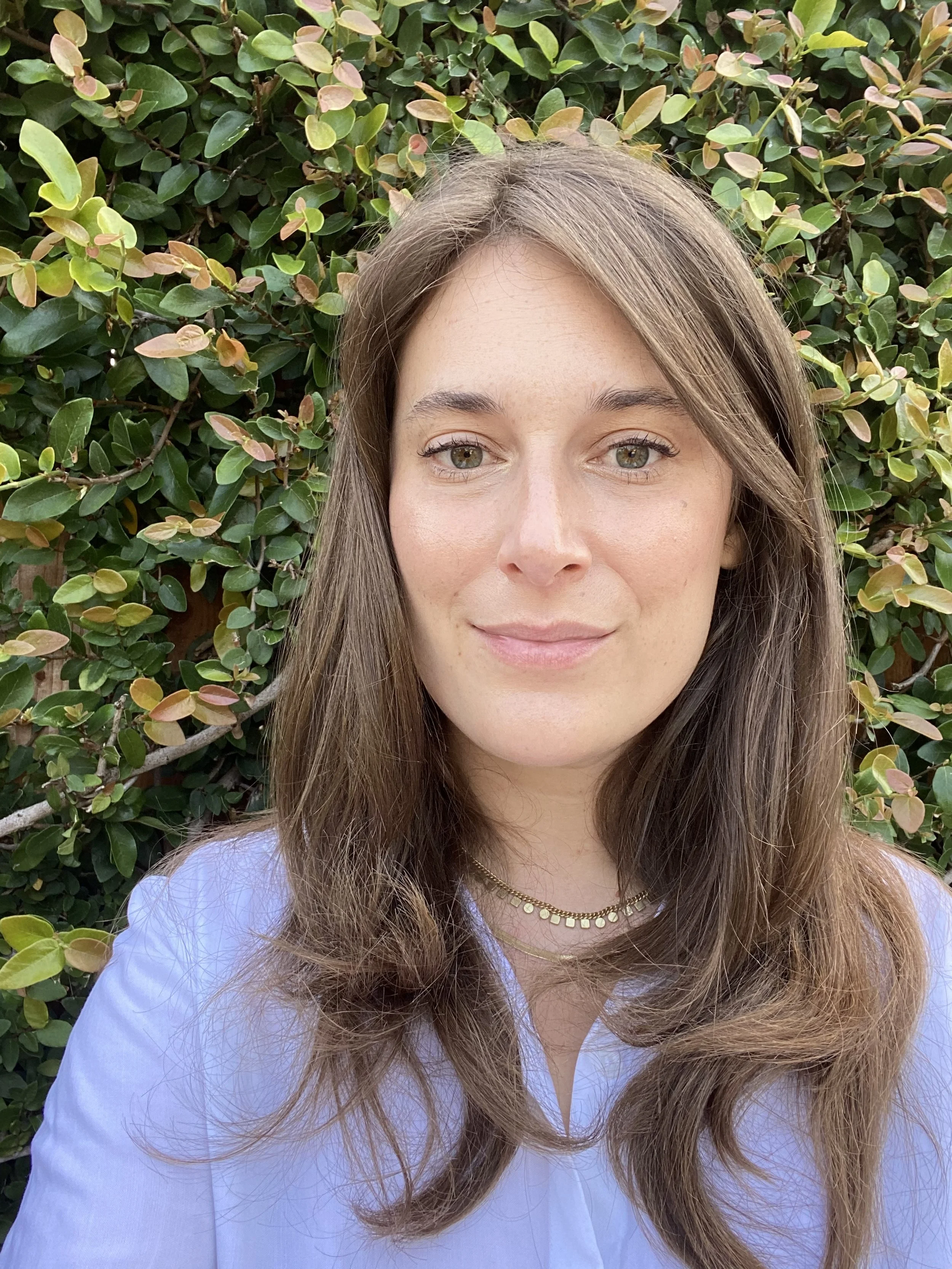 Portrait of a woman with long brown hair, light makeup, wearing a light blue shirt and gold necklace, standing outdoors in front of green leafy bushes.