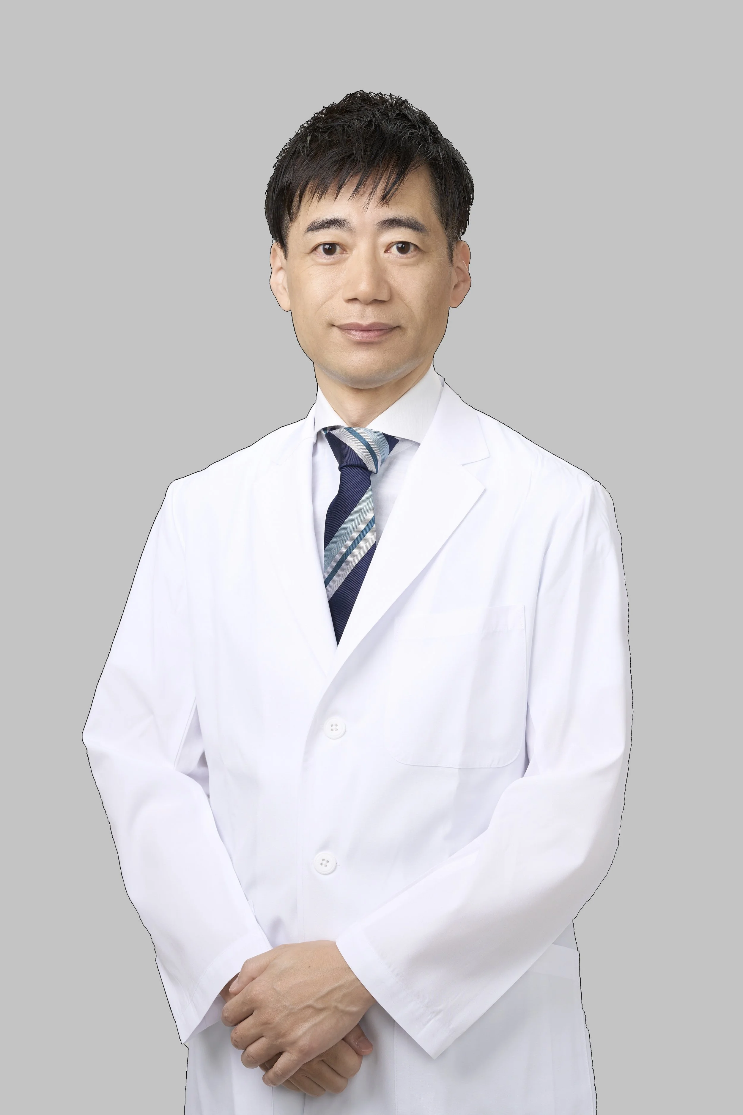 A male doctor wearing a white lab coat, tie, and shirt, standing against a plain gray background.