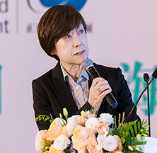 A woman with short dark hair speaking into a microphone at a table decorated with flowers.