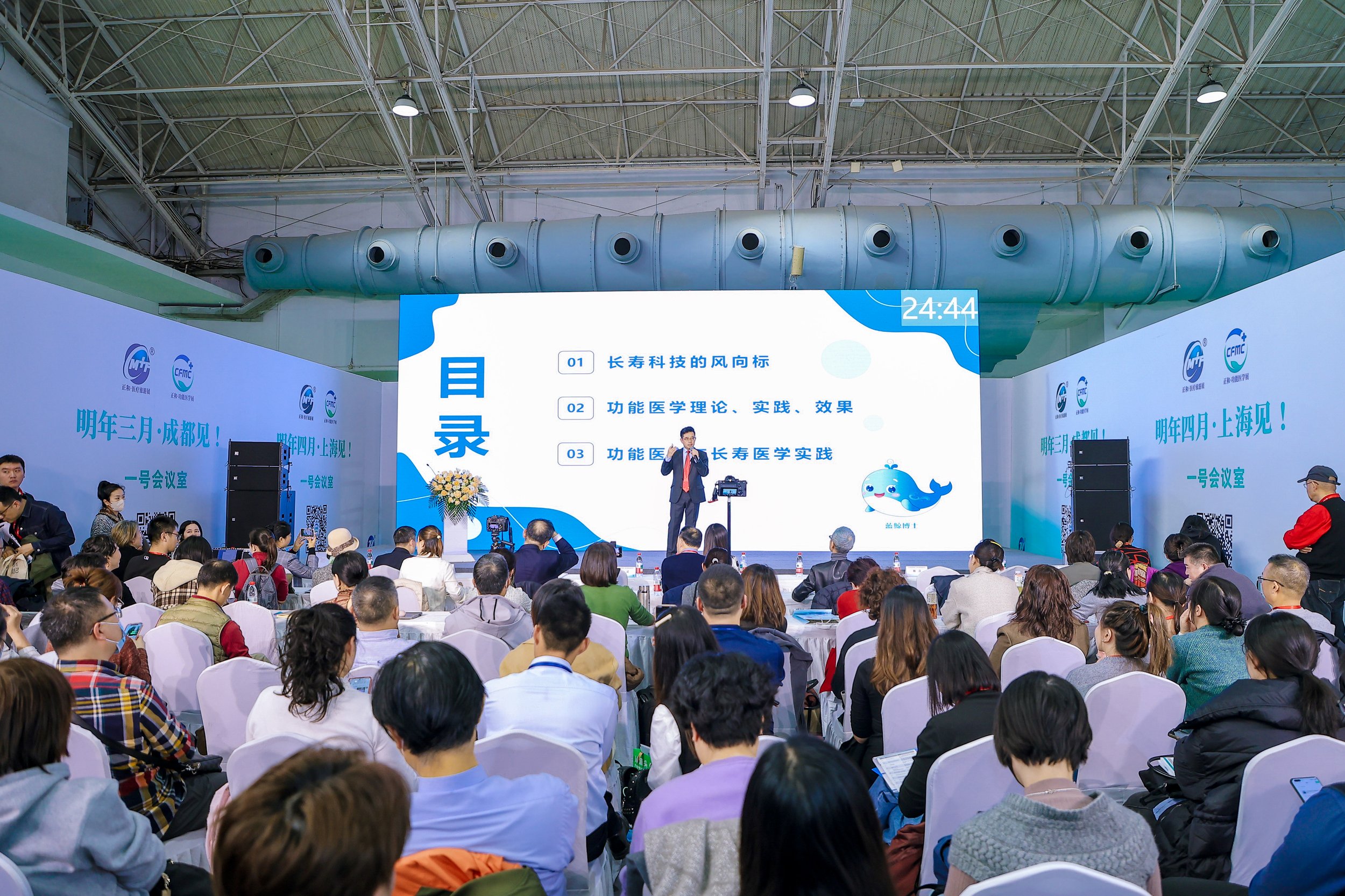 Conference room filled with seated audience watching a man give a presentation on stage. Large screen displays Chinese text and a cartoon whale. Stage is decorated with flowers, and banners in Chinese are visible on the sides of the room.