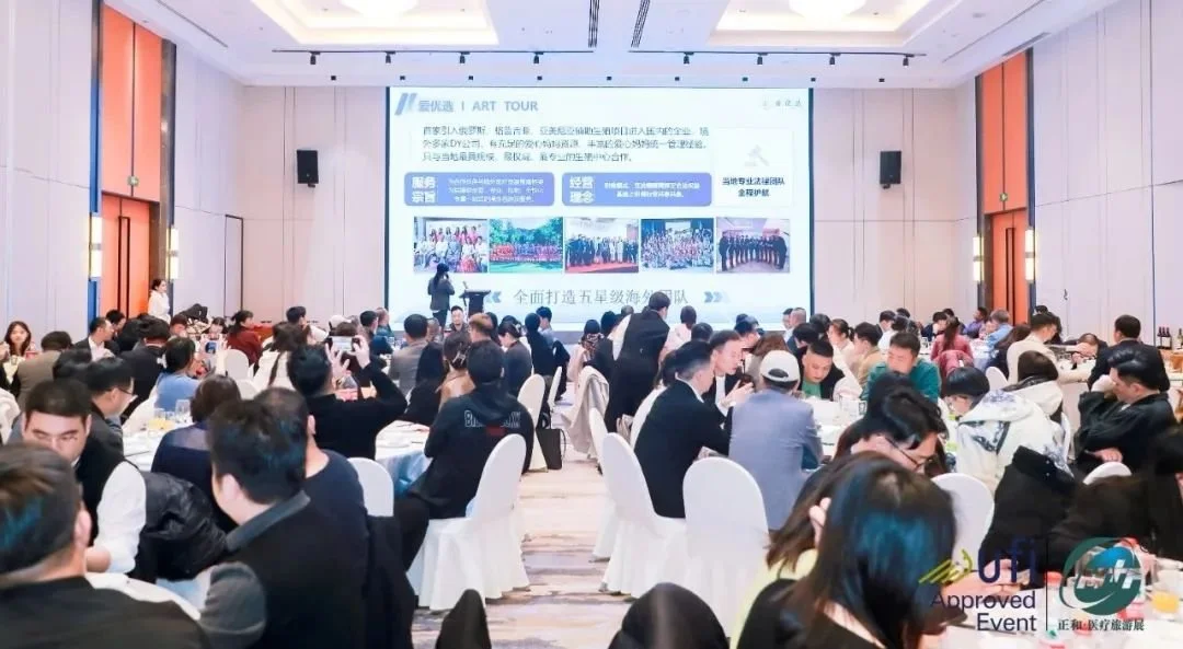 A large conference room filled with people attending a presentation or seminar. The audience is seated at round tables, listening to a speaker on stage with a large screen displaying a PowerPoint presentation in the background.