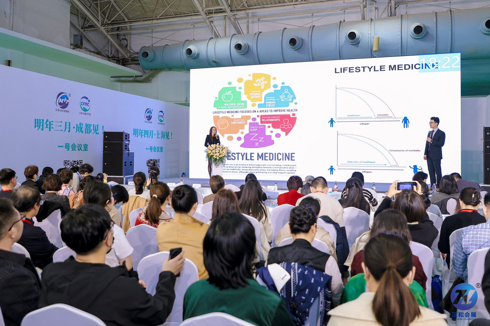 A presentation at a conference about lifestyle medicine features two speakers on stage in front of a large screen with charts and colorful graphics. The audience is seated and watching attentively.