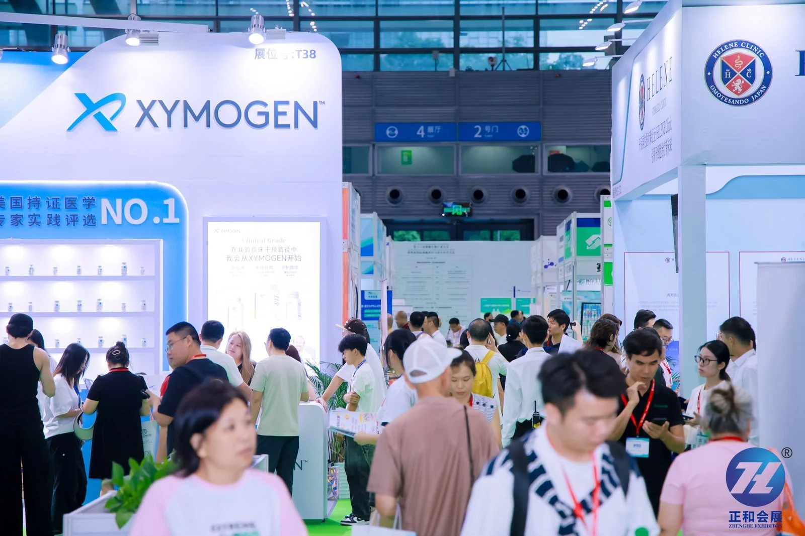 A busy exhibition hall with various booths and many visitors, with prominent signage for Xymogen and Helene Clinic.