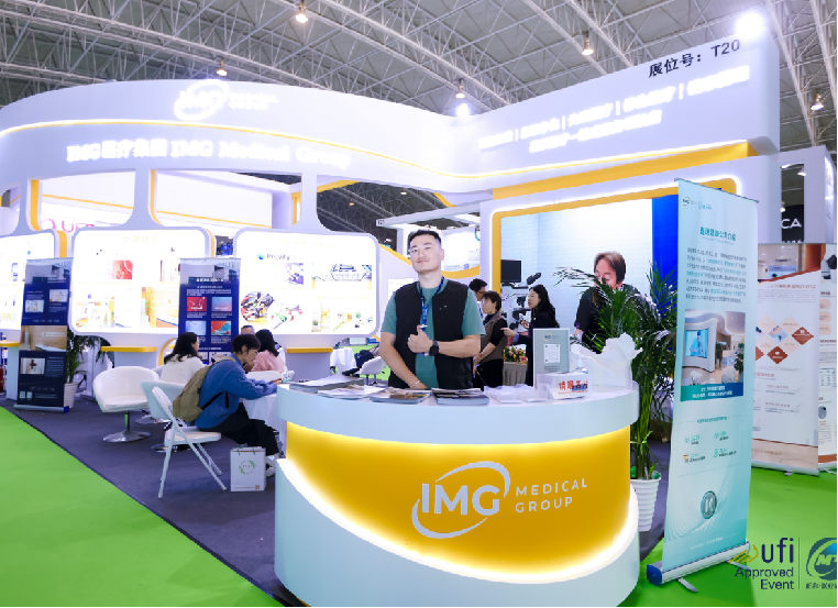 Trade show booth for IMC Medical Group with a curved yellow and white counter, company logo, and multiple informational posters. Several people, including staff and visitors, are interacting in a brightly lit, spacious exhibition hall with a green carpet.
