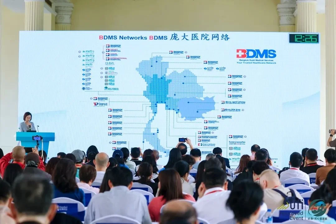 A large conference room filled with attendees watching a presentation on a giant screen displaying a map of Thailand with marked locations. A woman is speaking at a podium on the left side of the stage. The screen shows the logo for Bangkok Dust Medical Services (BDMS) and the title 'DMS Networks' in English and Chinese.