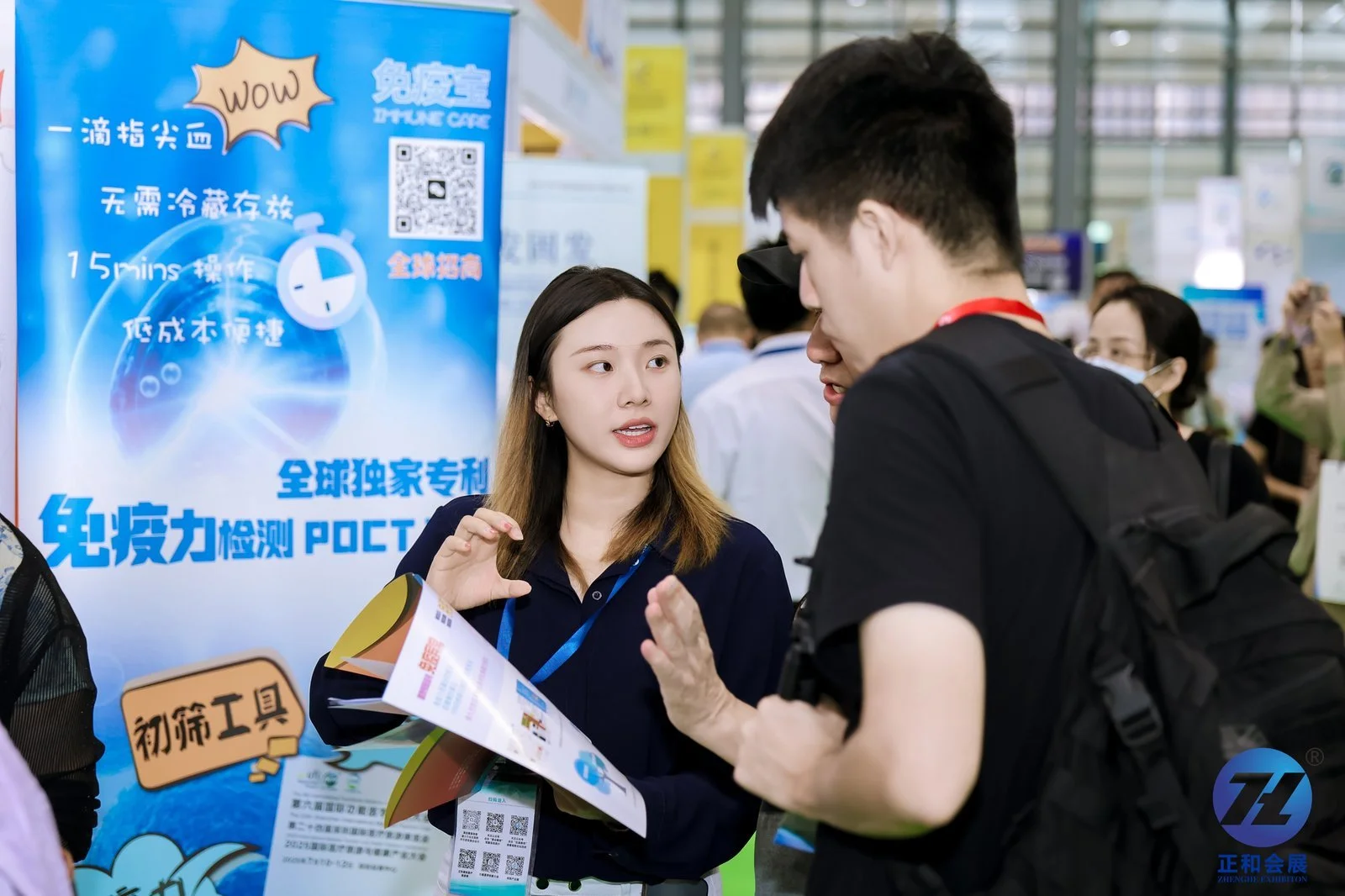 A woman with shoulder-length hair and a dark shirt is speaking to a man with a black backpack, wearing a black shirt, at a booth with a blue poster displaying Chinese characters, a QR code, and a clock graphic. The woman is holding a flyer and appears to be explaining something to the man. There are other people in the background, some wearing face masks, at an indoor event or exhibition.