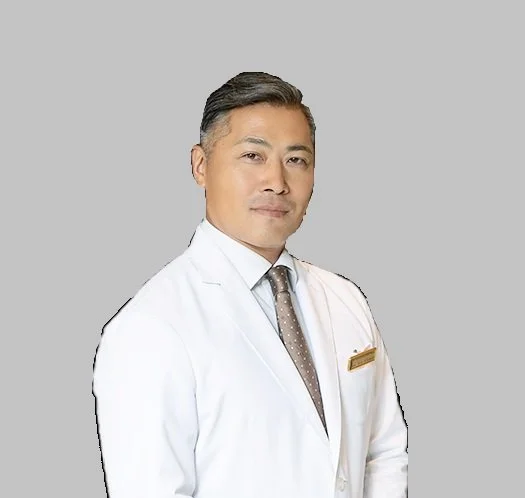 A male doctor with gray hair, wearing a white coat and a gray tie, standing against a plain background.