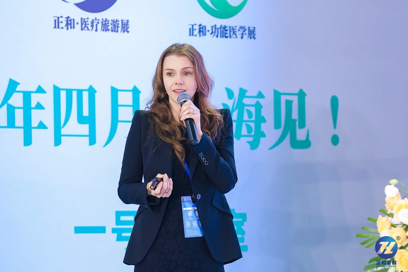 A woman with long brown hair wearing a dark blazer and dress giving a presentation with a microphone in front of a blue and green banner with Chinese characters and the ZL logo.