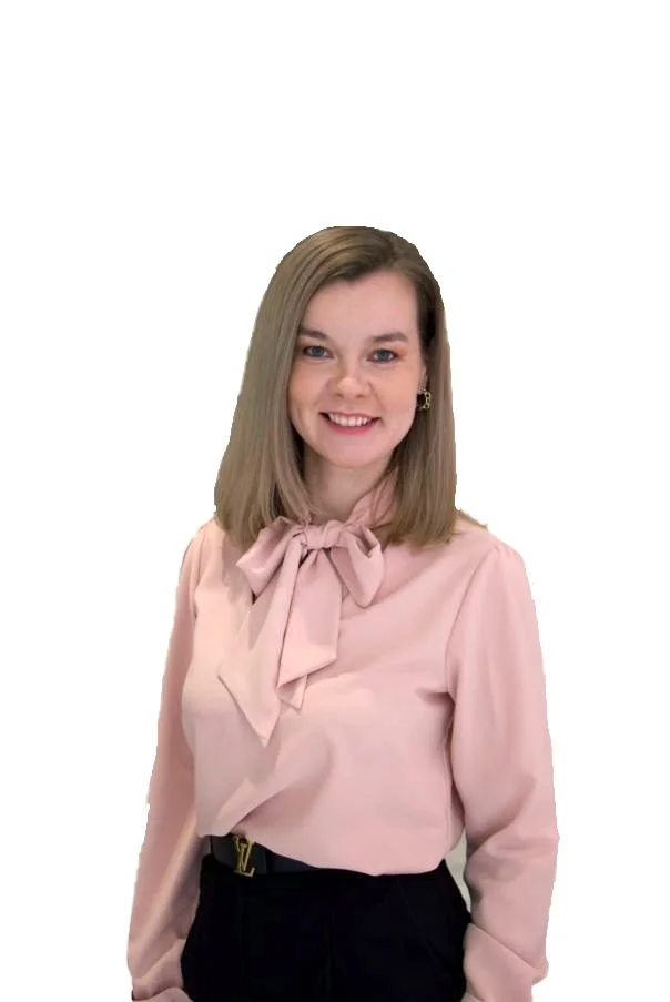 A woman with shoulder-length blonde hair, wearing a light pink blouse with a bow tie, and black pants, smiling at the camera against a plain white background.