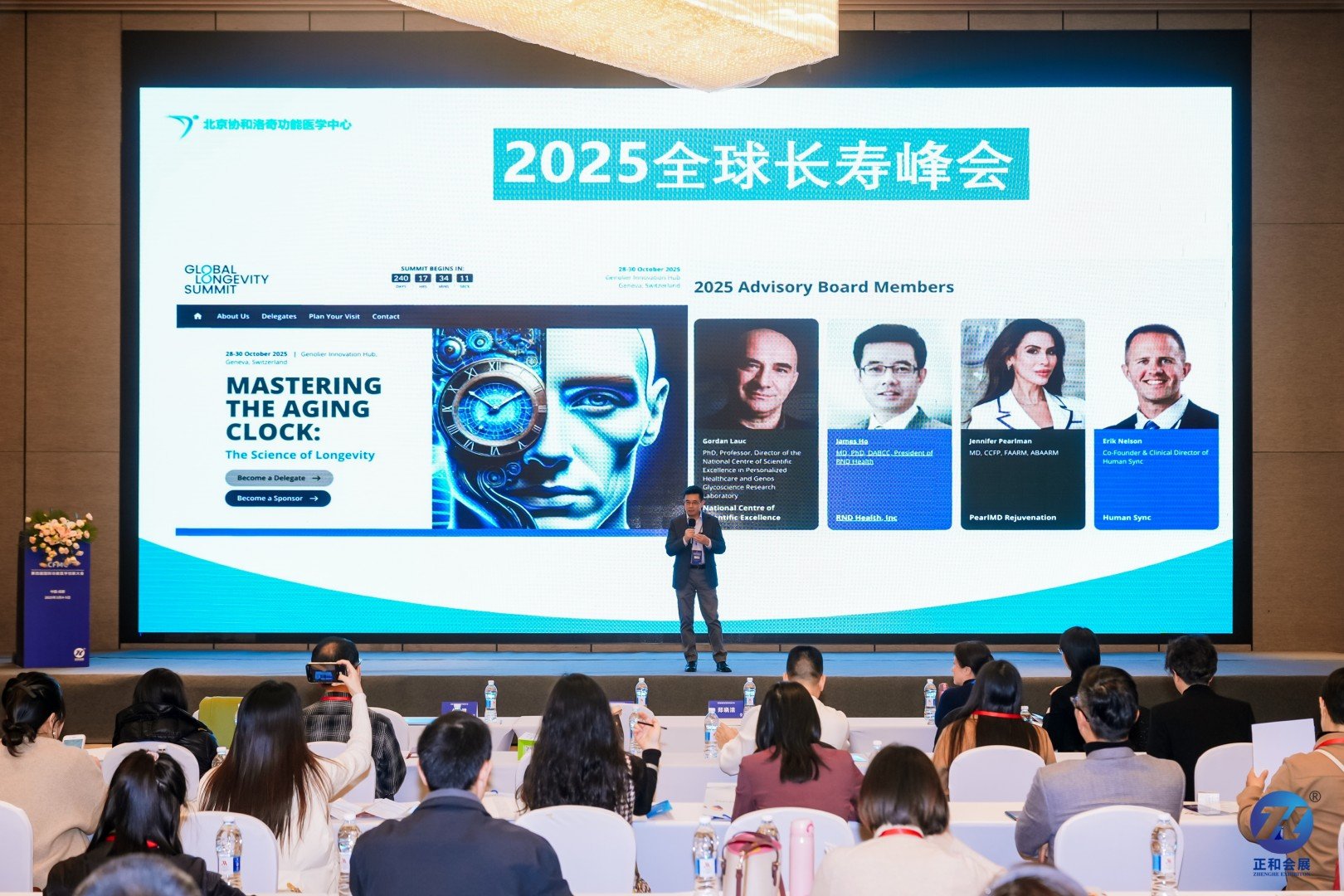 A conference room with attendees seated at tables facing a large screen displaying a presentation for the 2025 Global Longevity Summit, featuring speakers and event details, with a man standing on stage speaking to the audience.