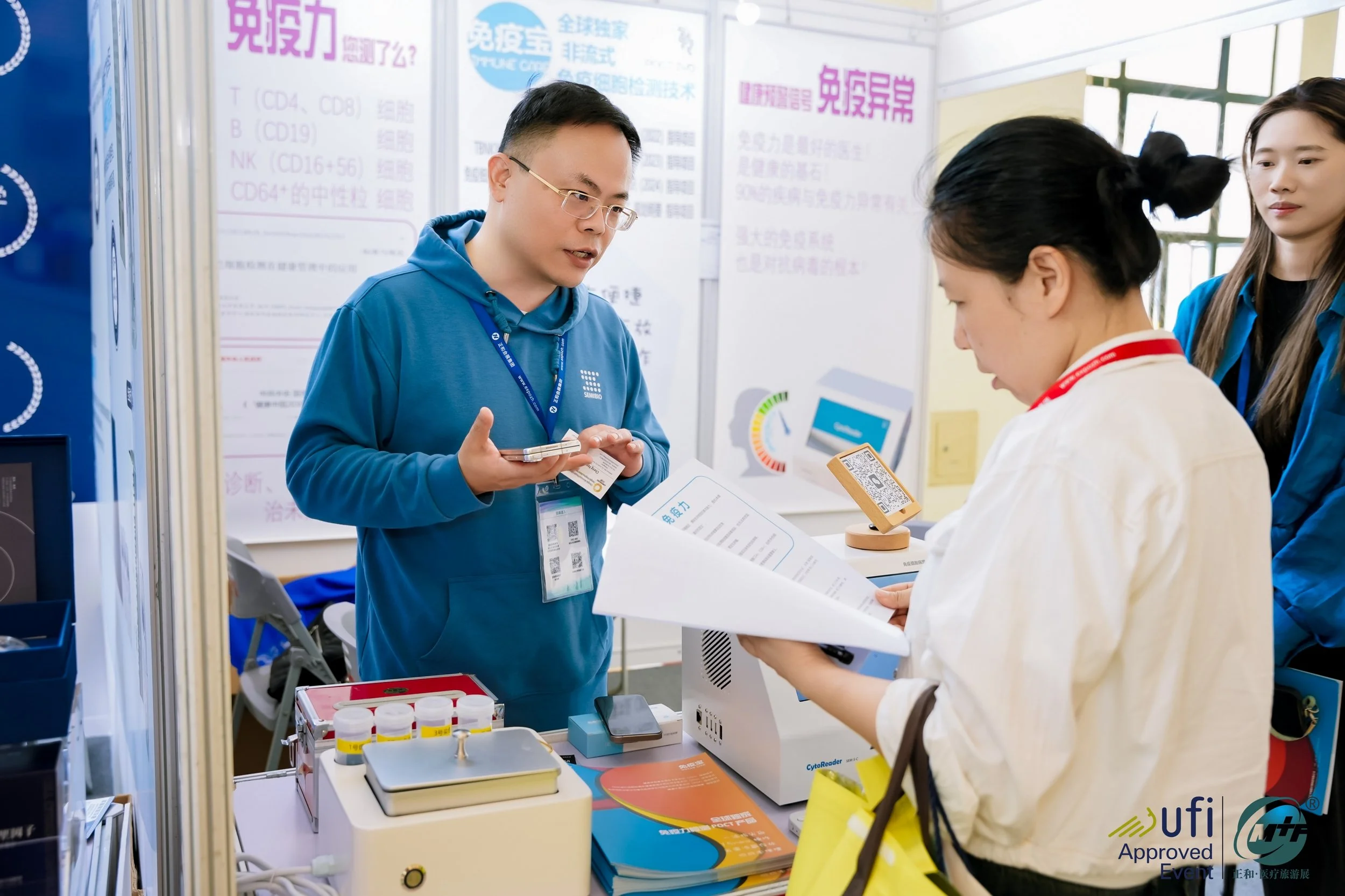 A man in a blue hoodie and glasses is talking to a woman in a white lab coat at a science exhibition booth, with informational posters in the background. The woman is holding a brochure, and there are scientific equipment and brochures on the table.