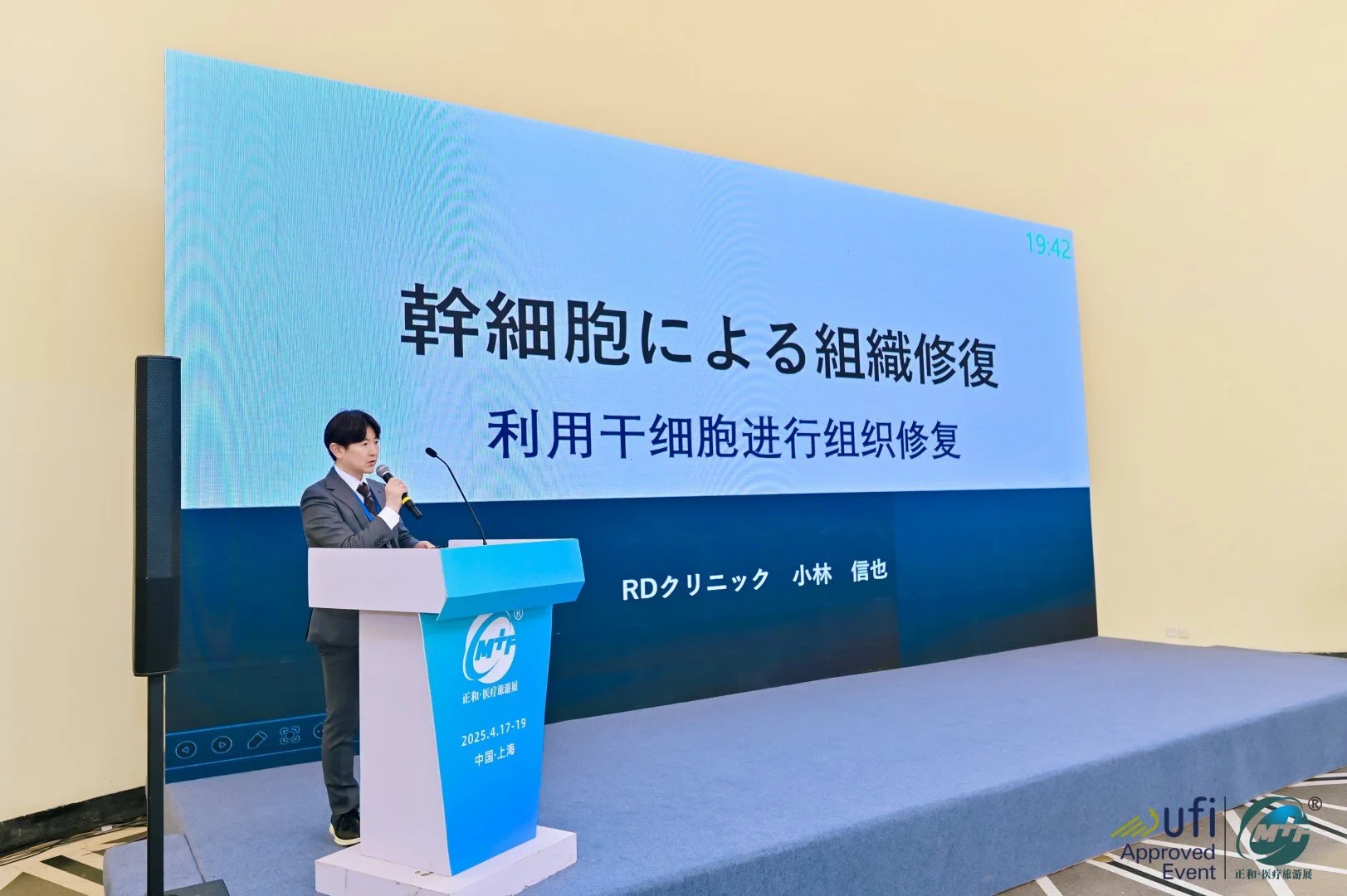 A man in a gray suit and blue tie standing at a podium holding a microphone, giving a presentation at a conference in Shanghai, China, with a large screen behind him displaying Japanese and Chinese text about stem cell tissue regeneration.