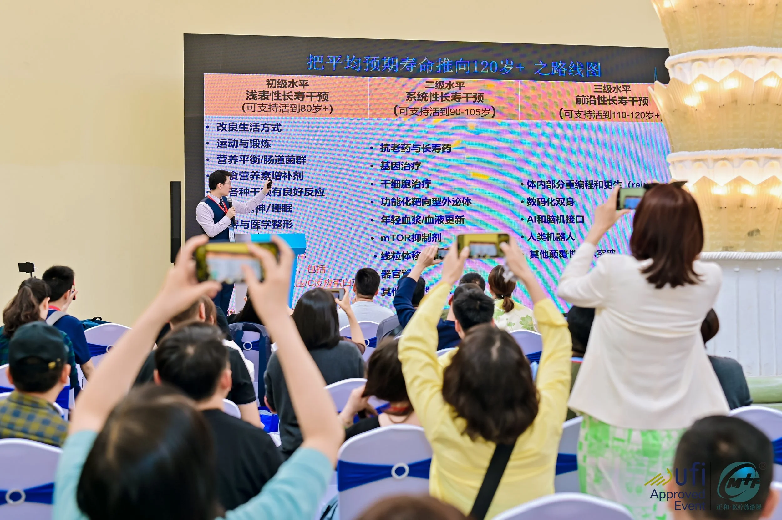 A presenter stands on stage giving a presentation to an audience seated in chairs, with some audience members recording or taking photos of the presentation on their phones. The large screen displays text in Chinese.