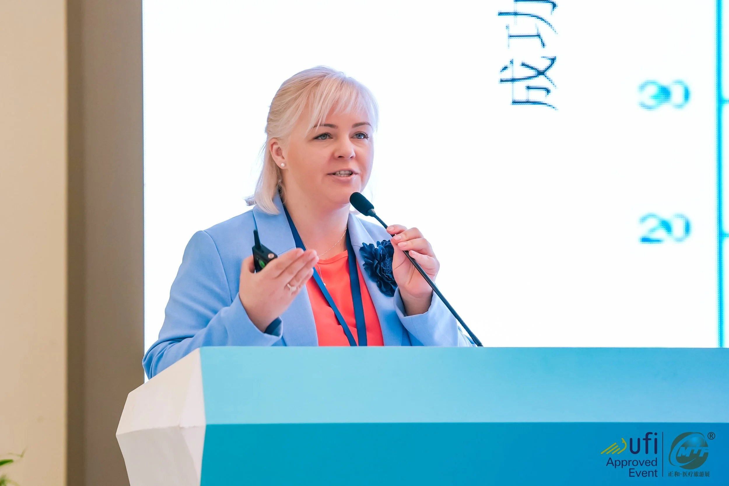 A woman giving a presentation at a conference, standing behind a light blue and white podium, holding a remote control, and speaking into a microphone. She has blonde hair, wears a blue blazer with a dark blue flower pin, and a coral top. There is a large screen behind her displaying a presentation with blue text and numbers.
