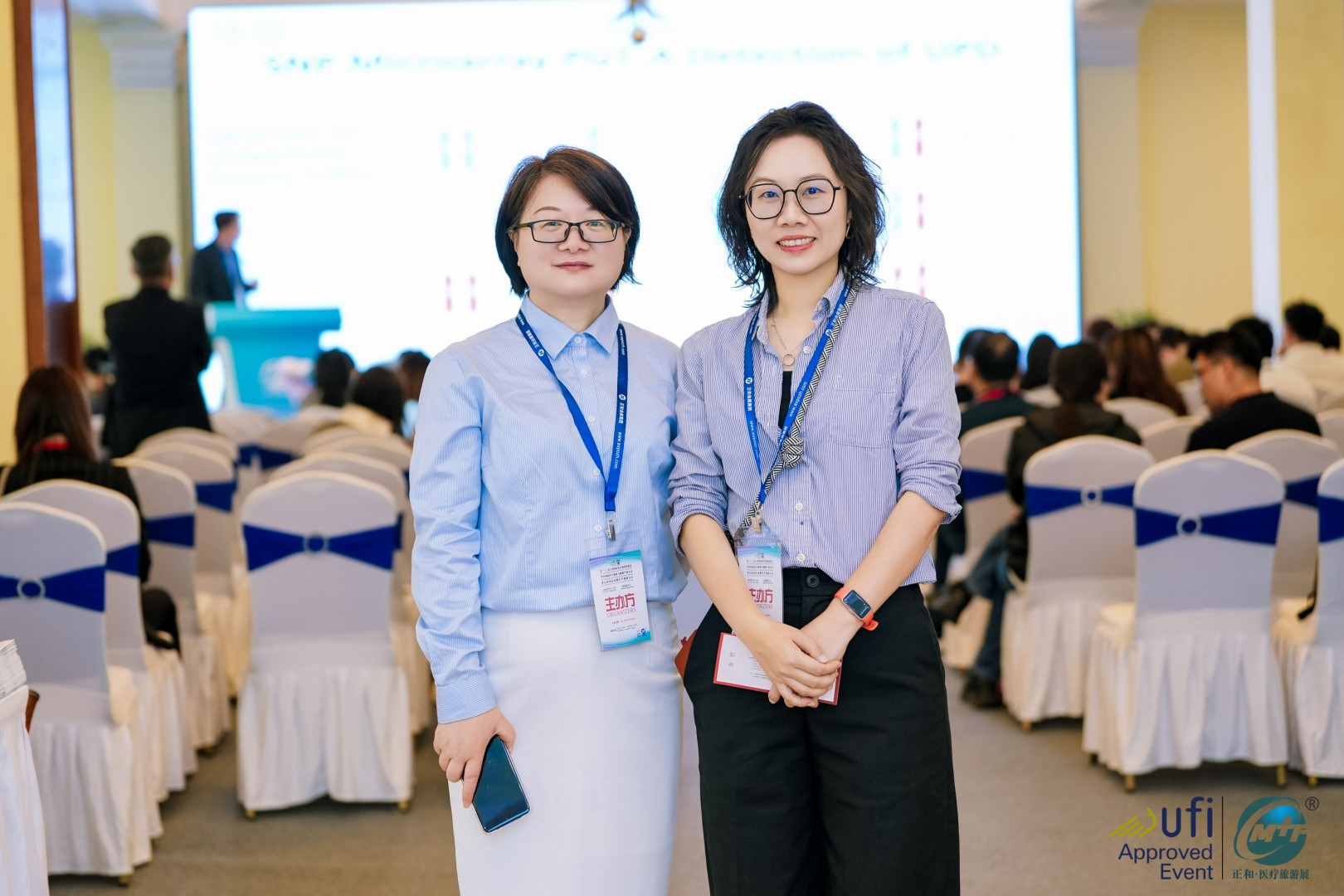 Two women with conference badges standing and smiling in a conference hall filled with seated attendees and a large presentation screen in the background.