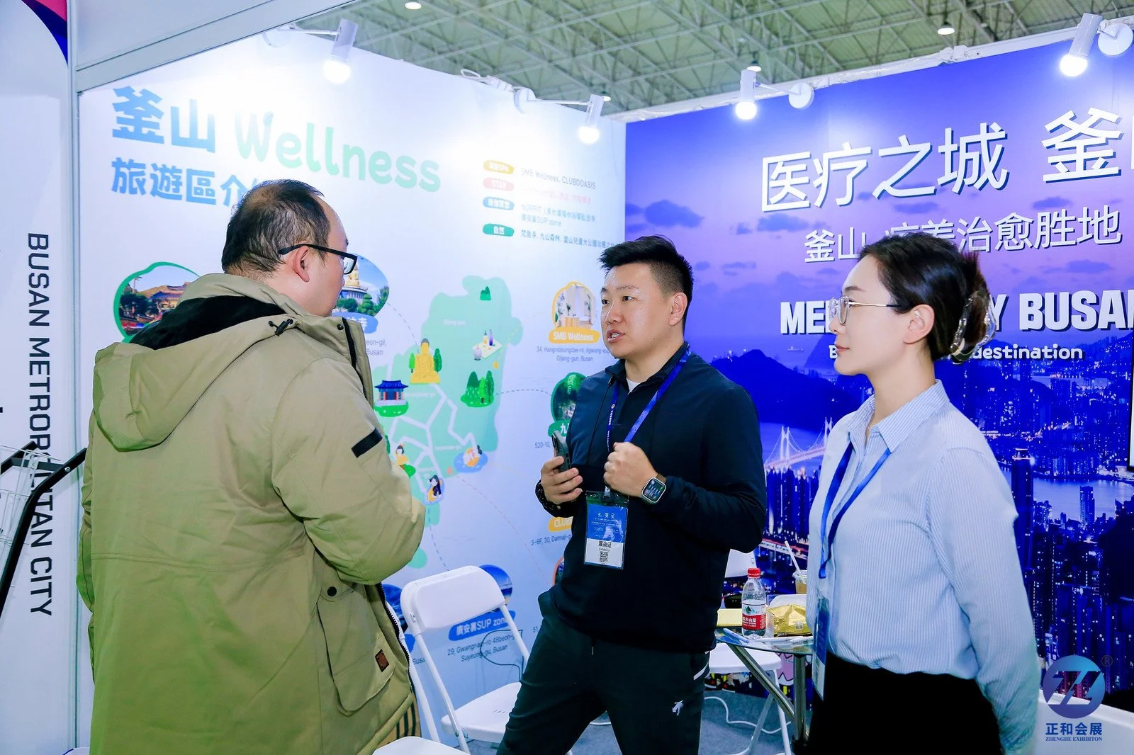 Three people having a conversation at a booth with promotional materials and a cityscape backdrop, promoting Busan, South Korea.