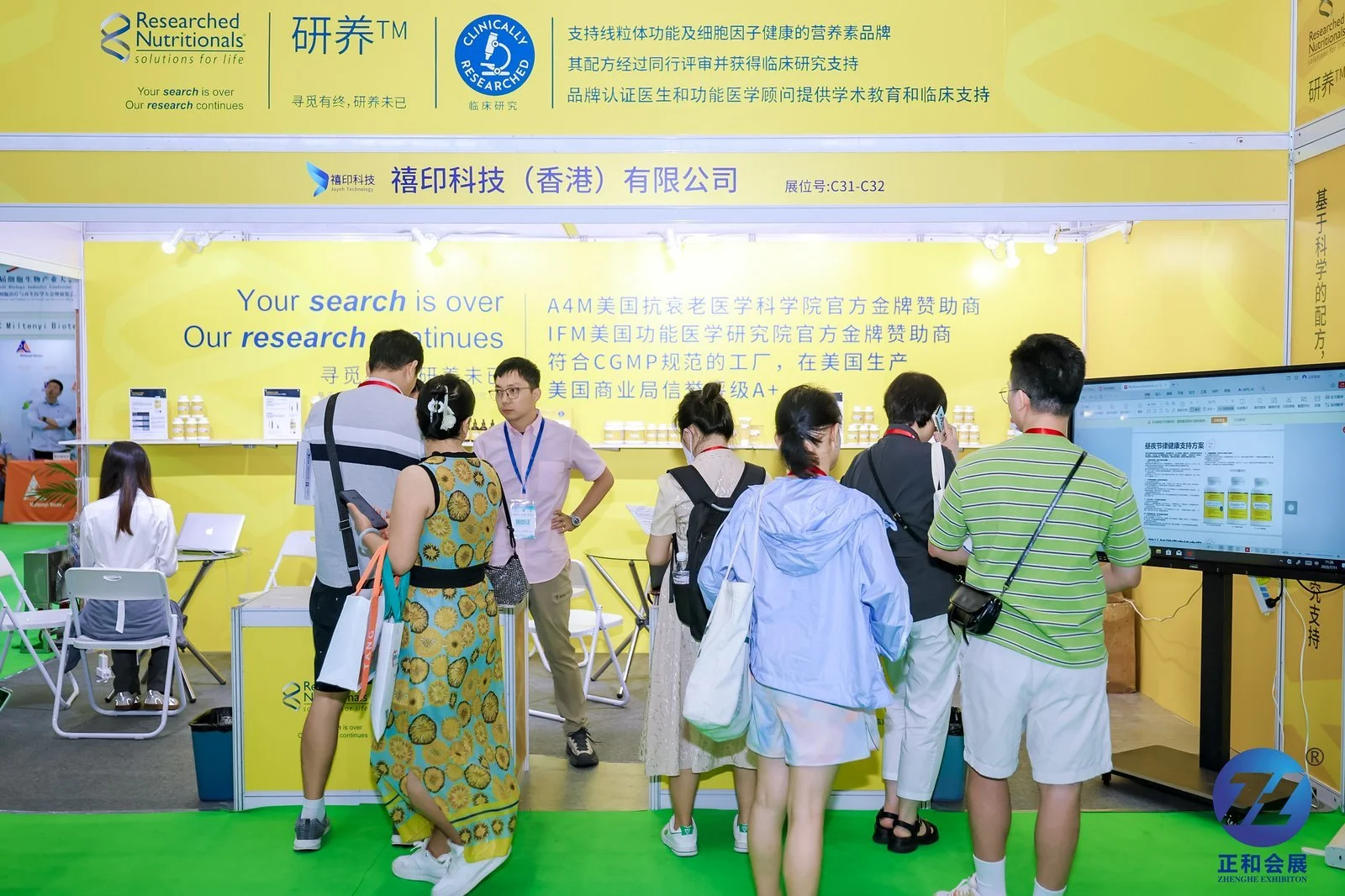 Exhibition booth for Researched Nutritionals at a trade show, with several visitors engaged and looking at product displays and a large screen showing information, all set against a bright yellow background with business information and promotional messages.