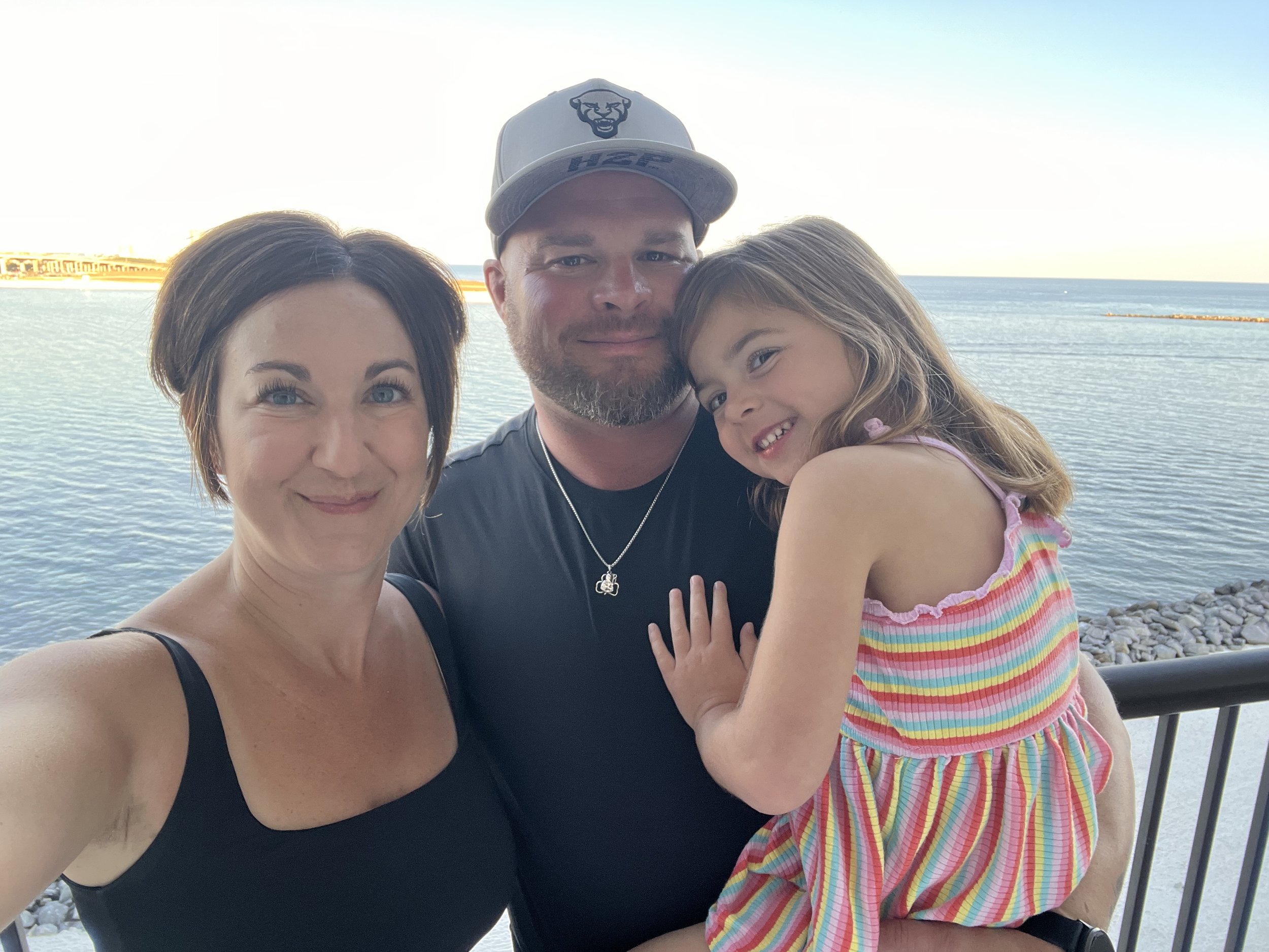 A family of three taking a selfie outdoors near the water, with the ocean and rocky shoreline in the background. The woman has short brown hair and is wearing a black top, the man is wearing a black shirt and a baseball cap, and the young girl has shoulder-length light brown hair and is wearing a colorful striped dress.