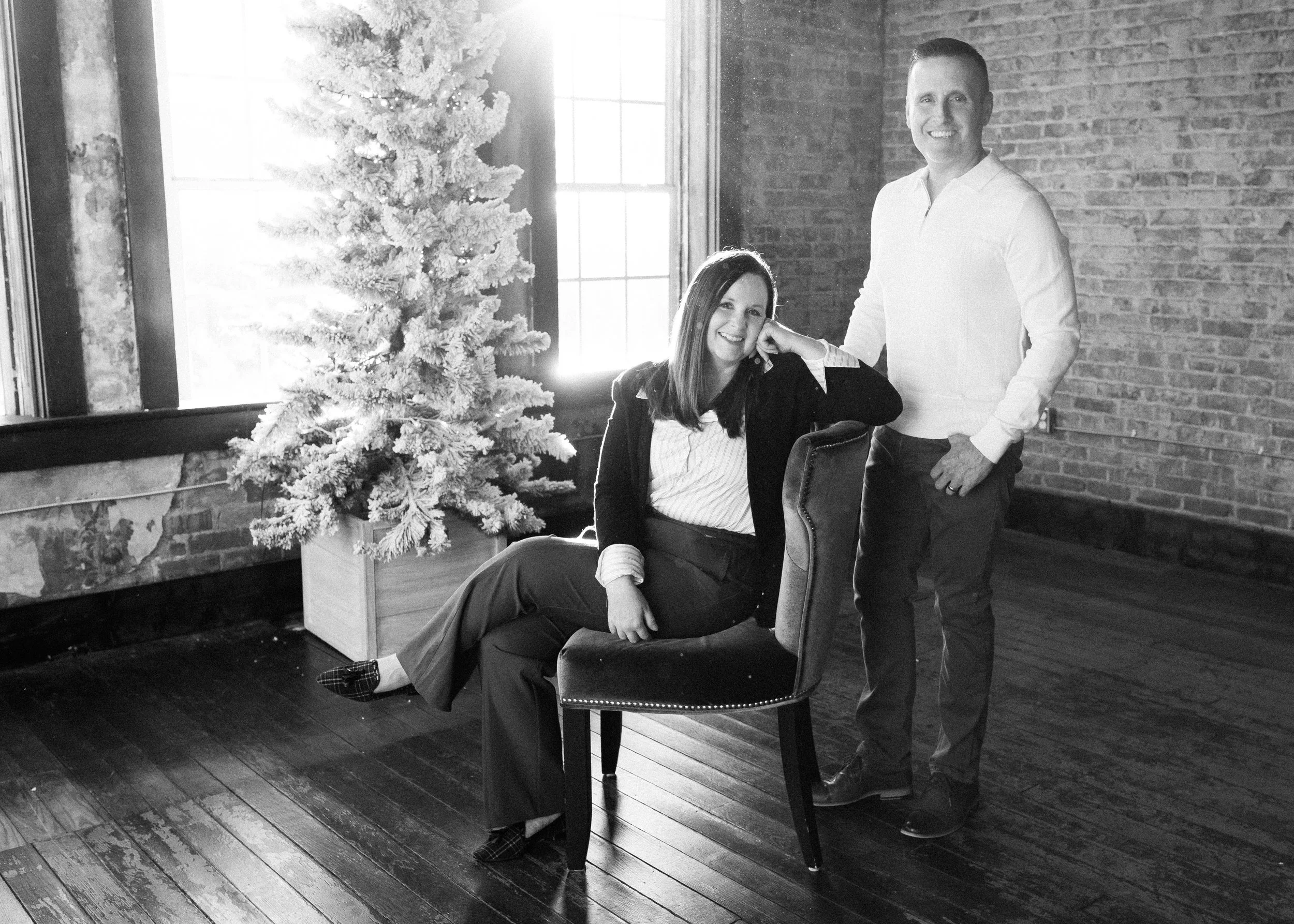 A woman sits on a chair with her legs crossed, resting her head on her hand and smiling, while a man stands beside her, smiling at the camera. There is a decorated Christmas tree and a window in the background with sunlight shining through. The setting has exposed brick walls and wooden floors.