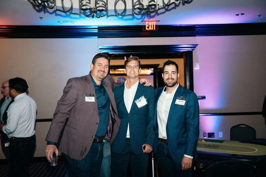 Three men in suits smiling at a business event, one with a cane, in a conference room with purple and blue lighting and an EXIT sign overhead.