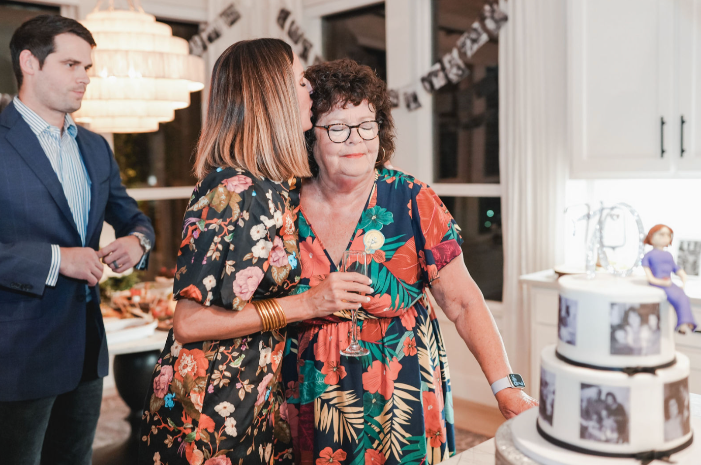 A woman in a floral dress holding a champagne flute is hugging a woman in a multicolored tropical dress at a celebration, with a decorated cake and photographs in the background.